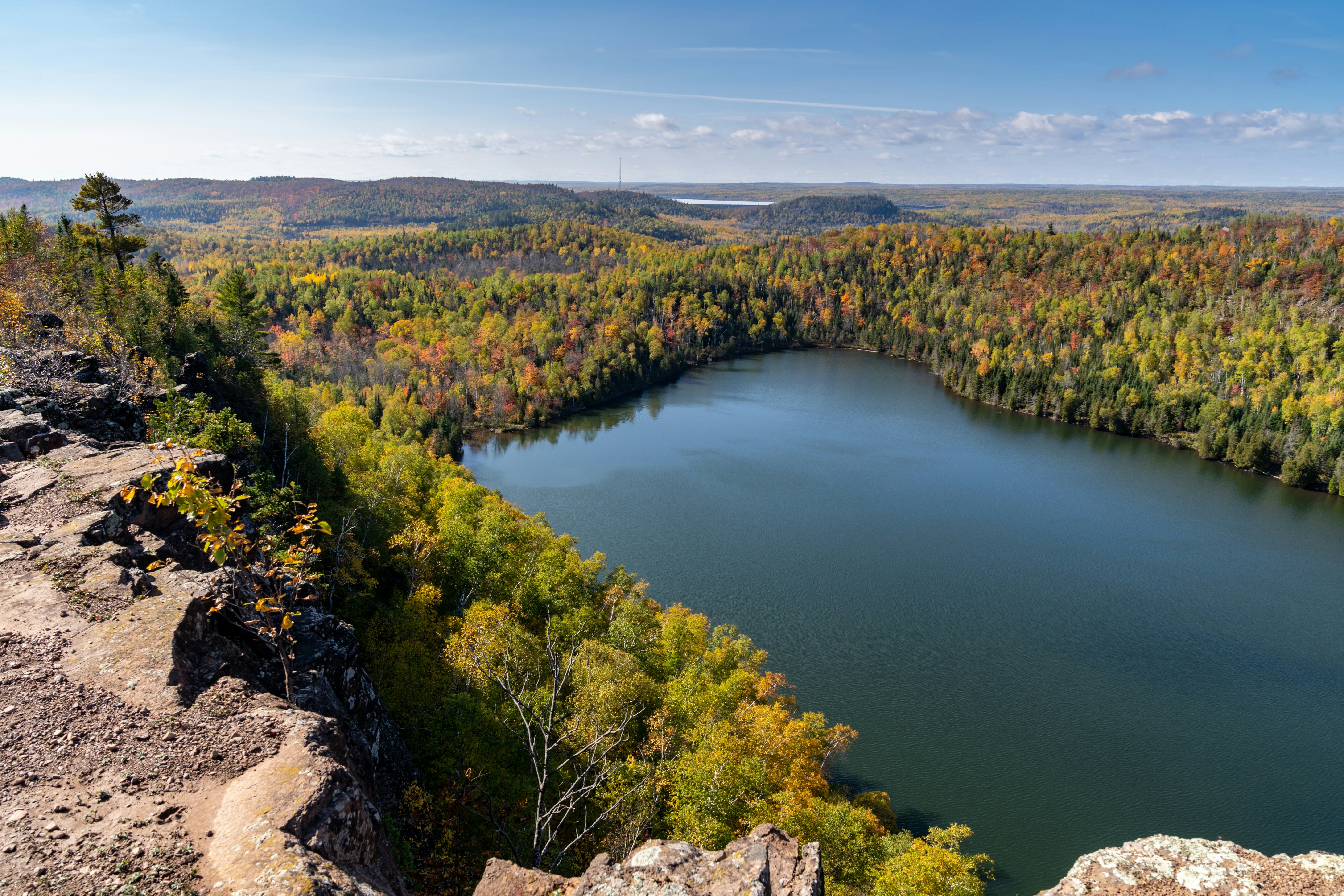 From a rocky outcrop, trees with some autumn color descend to the shore of a calm lake; the forest continues to the horizon, and another lake is in the far distance.