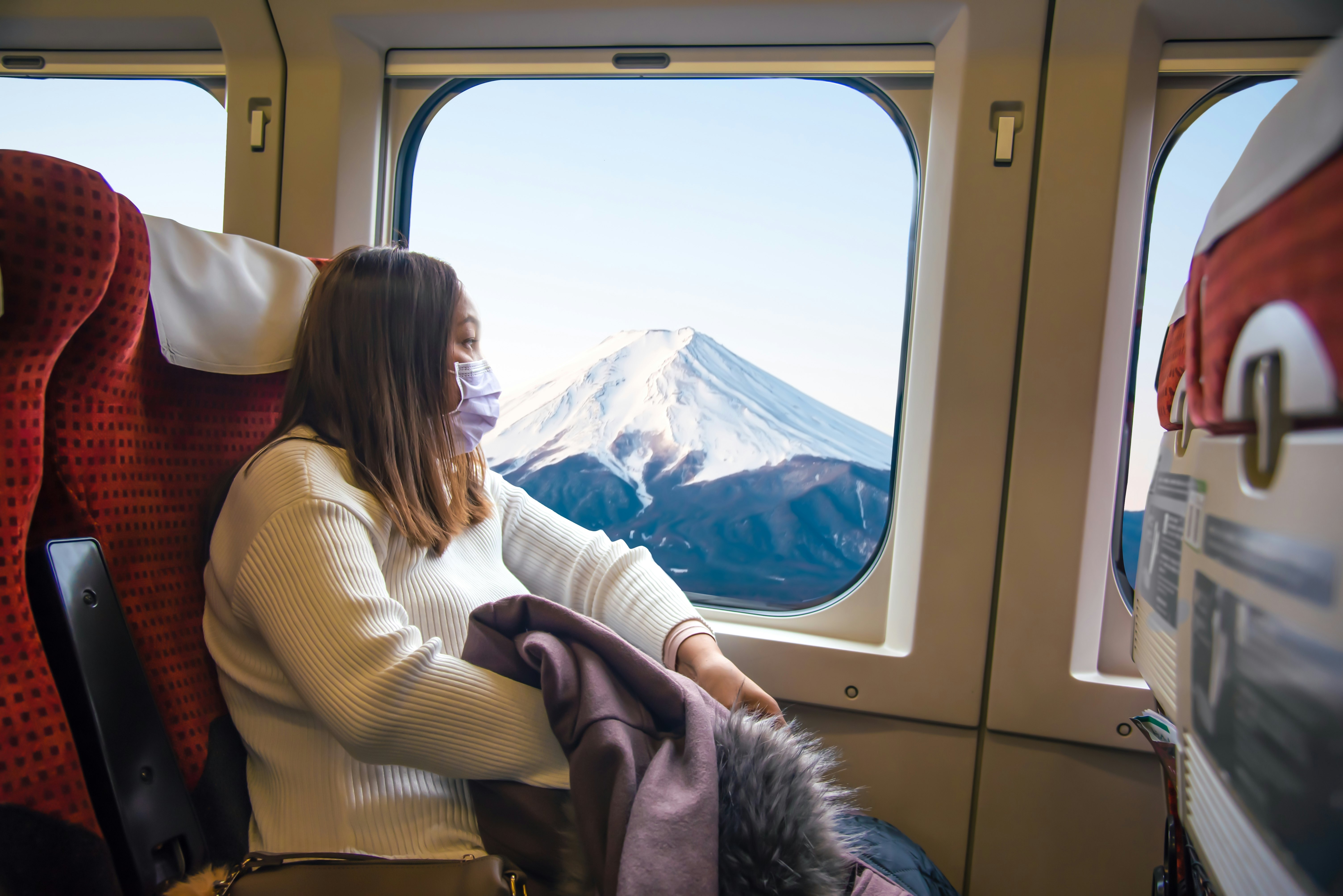 A masked young Asian woman sitting on a train. She is holding her purple coat in her lap and looking out a window where a snow-capped mountain is visible.