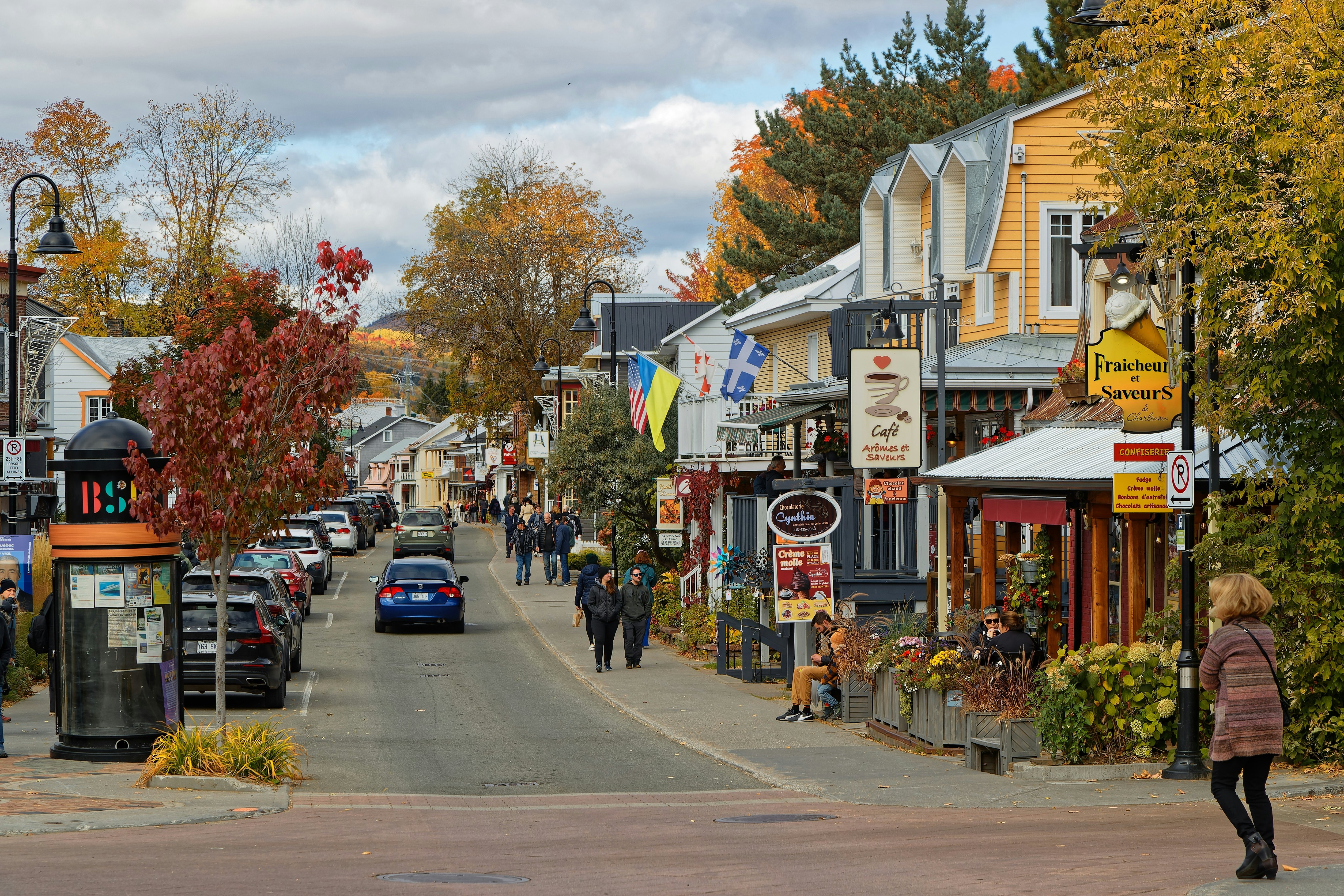 Cars drive away on a single-lane road that runs through a town; shops with signs and flags are on one side of the street, parked cards are on the other. The trees are in autumn colors, and people are walking on the sidewalk.