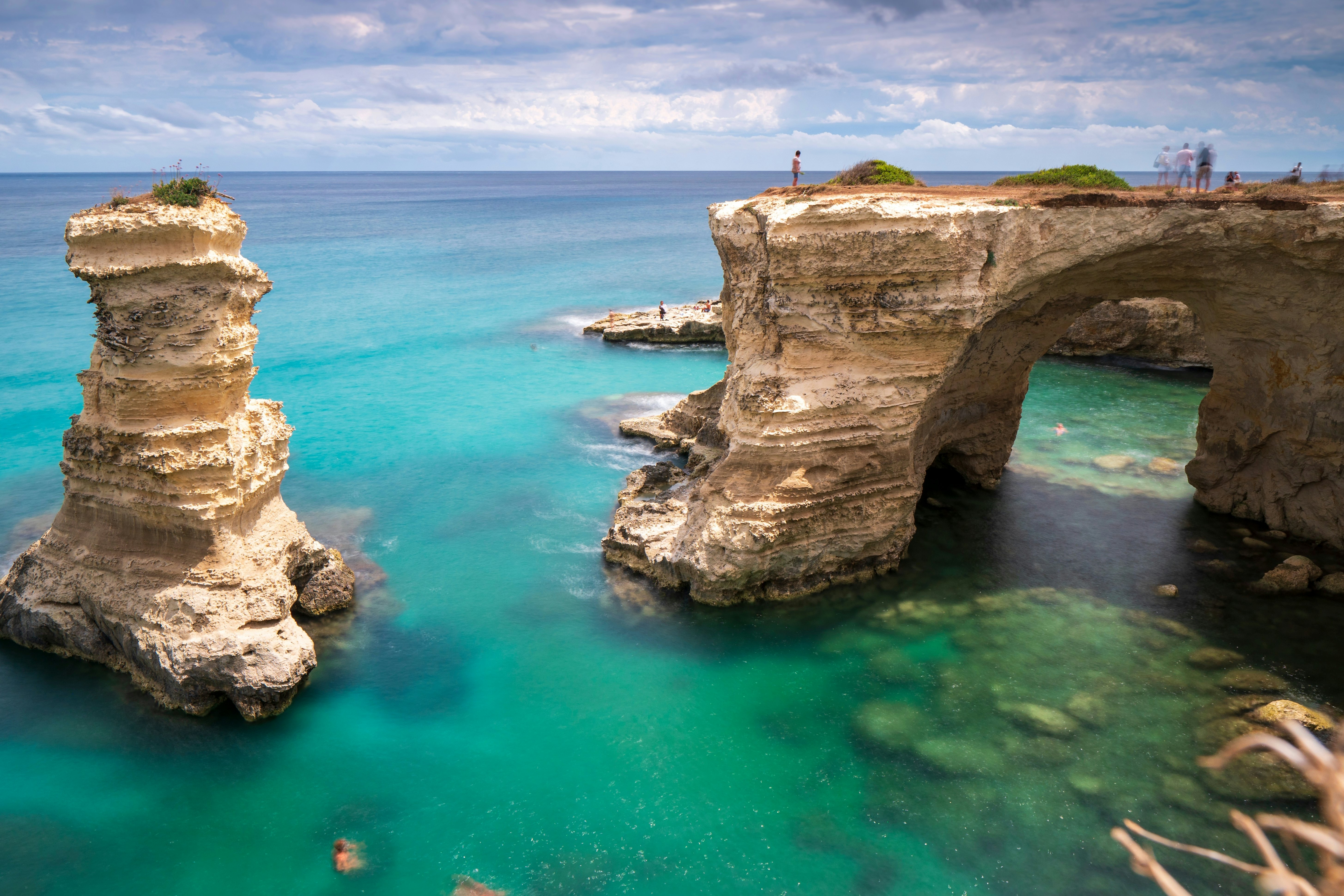 People stand on a vast natural rock arch out at sea, while others swim close to nearby rock stacks.