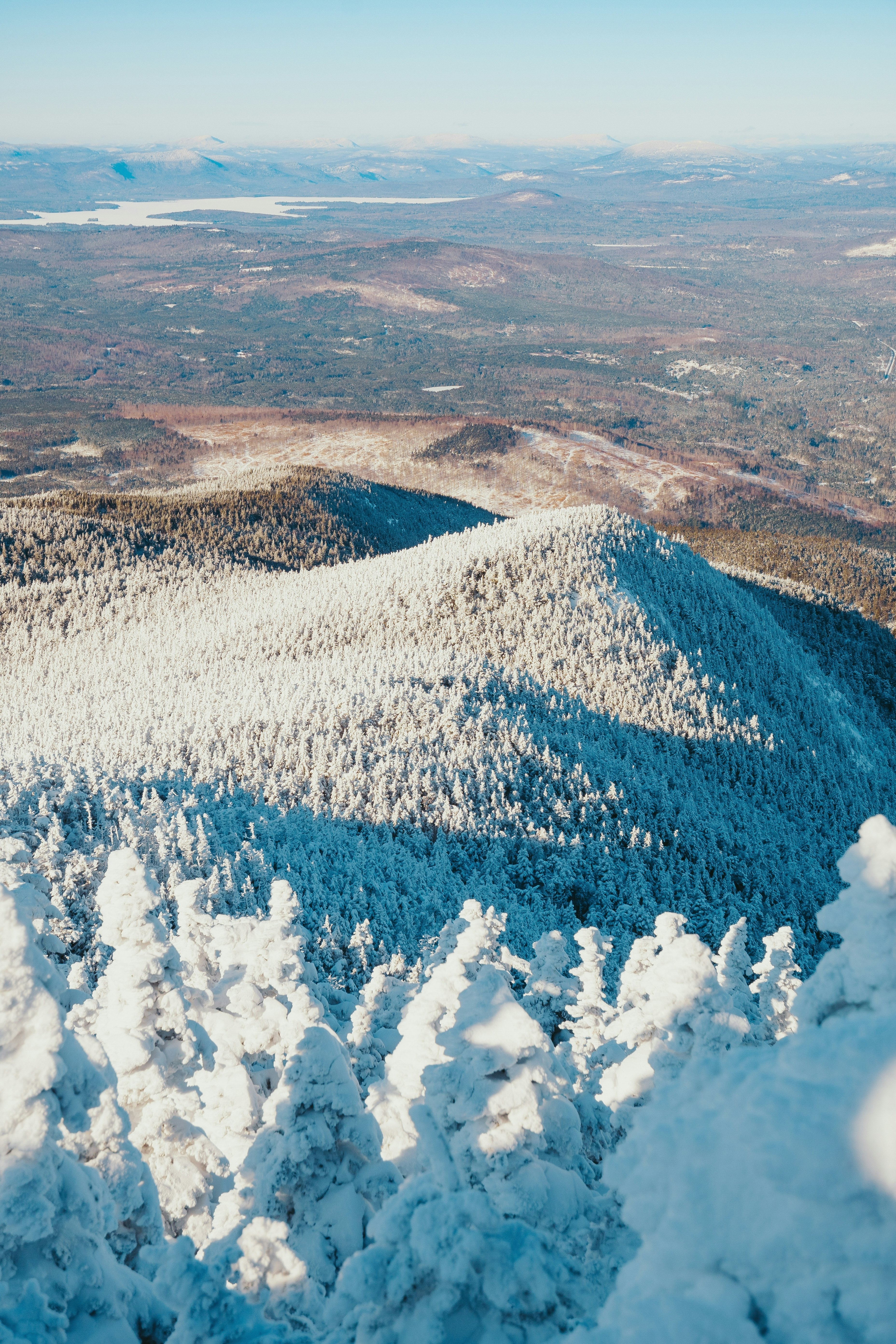 From the top of a mountain in Maine, snow-covered evergreen trees extend down to a valley with no snow.