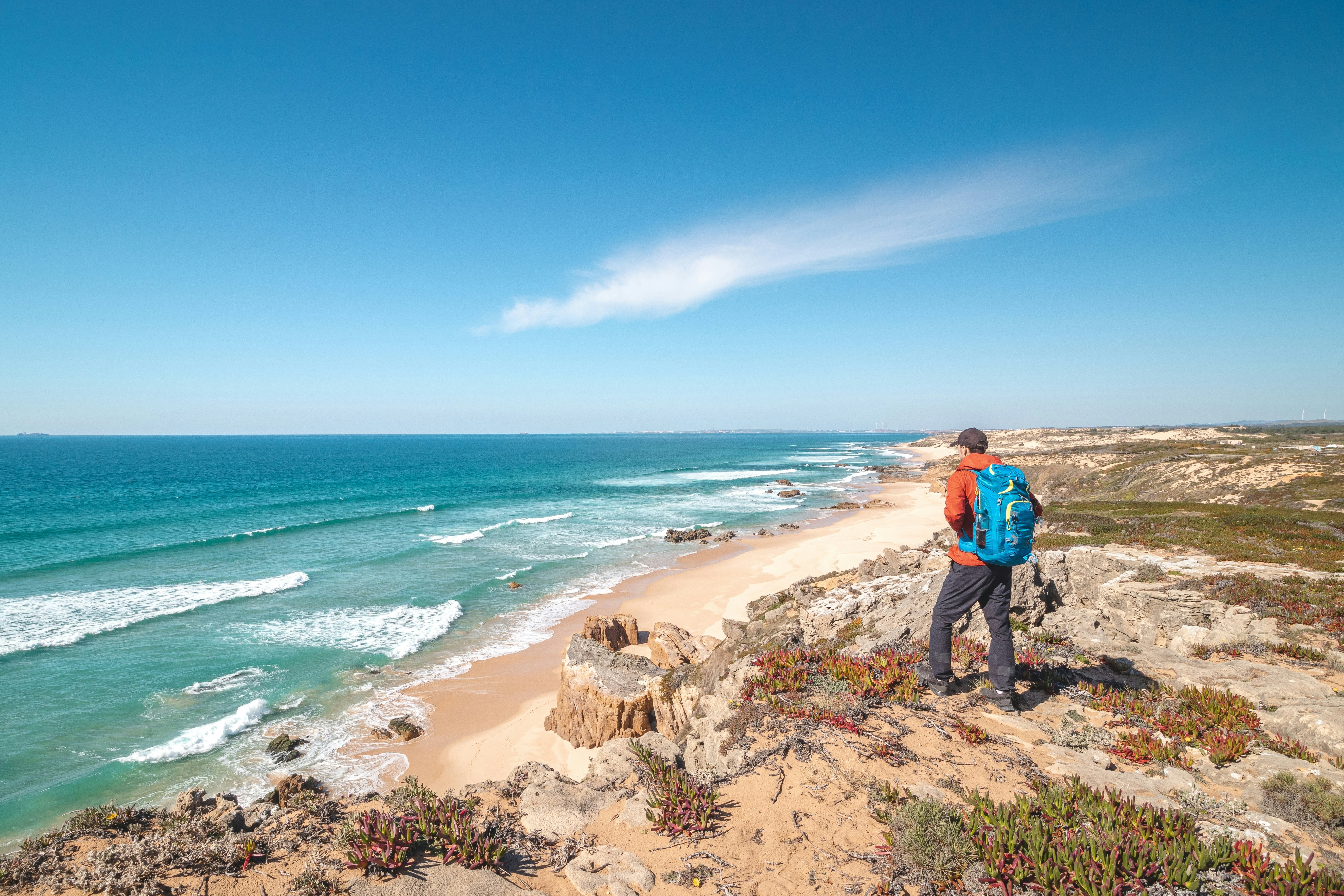 A hiker with a pack is seen from behind standing on dunes and looking out at the waves lapping a beach.