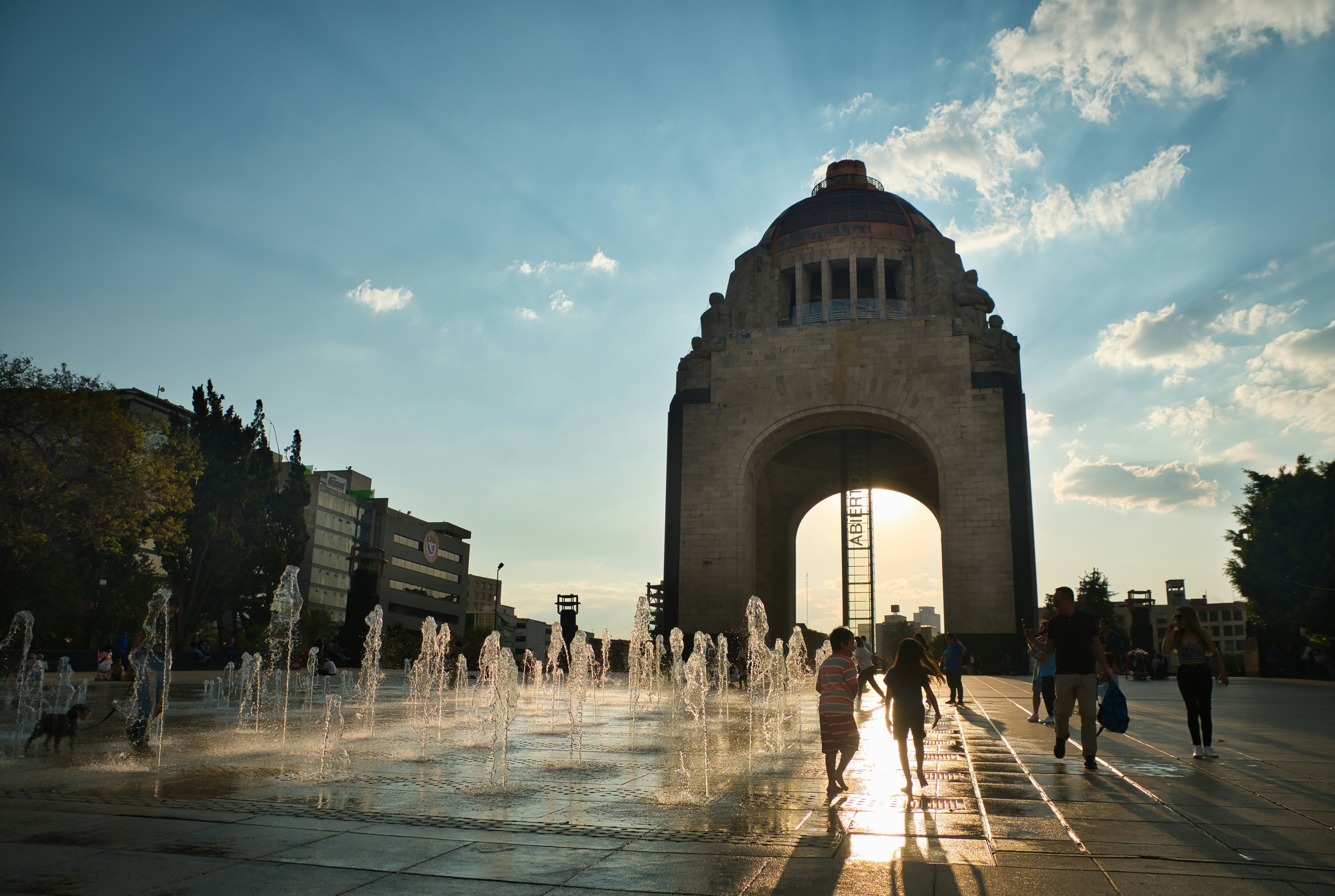 People run through a water feature on a plaza at dusk, with a huge monument seen at one end of the plaza.