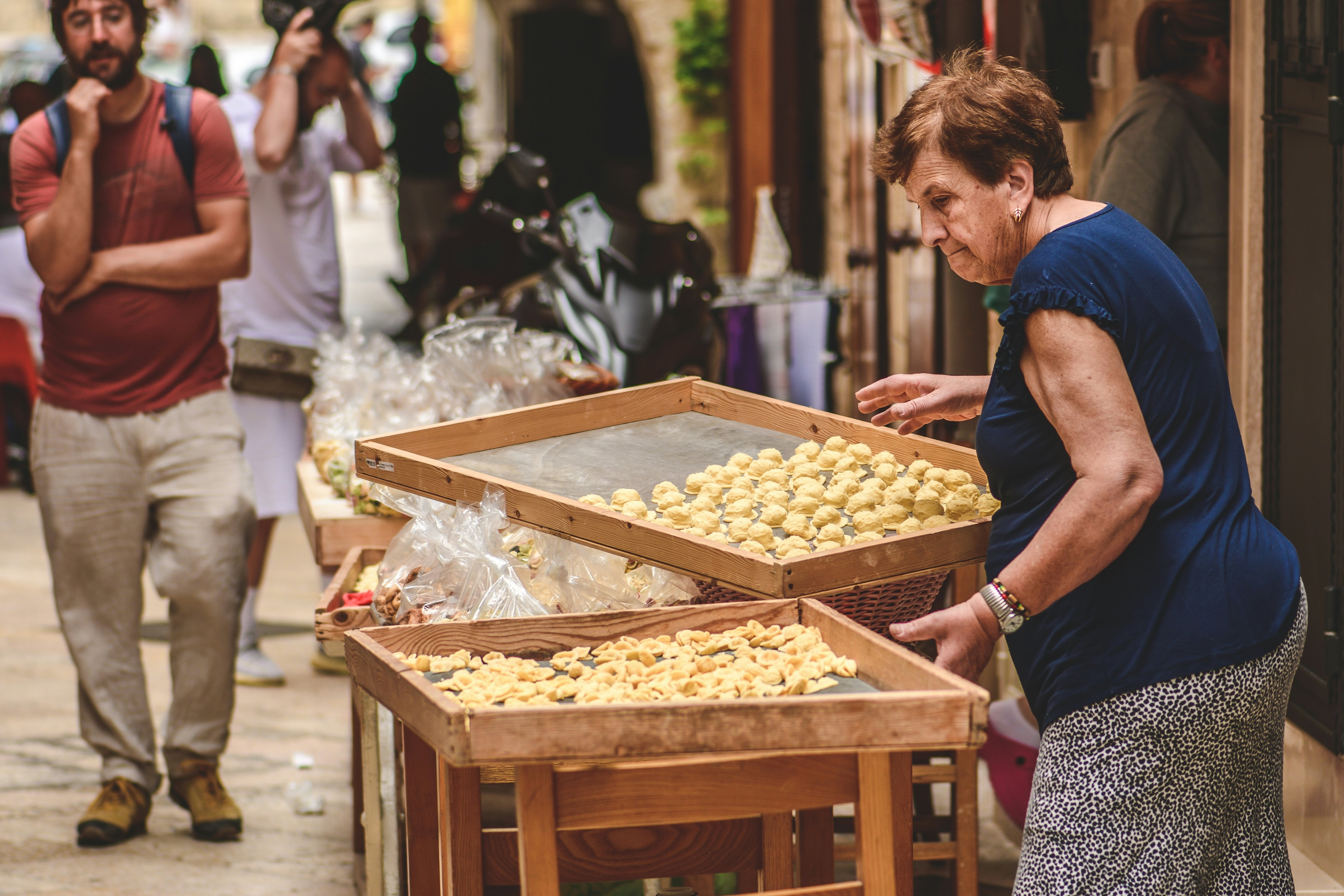 A senior woman sets out trays of freshly made pasta on tables in a narrow city street.