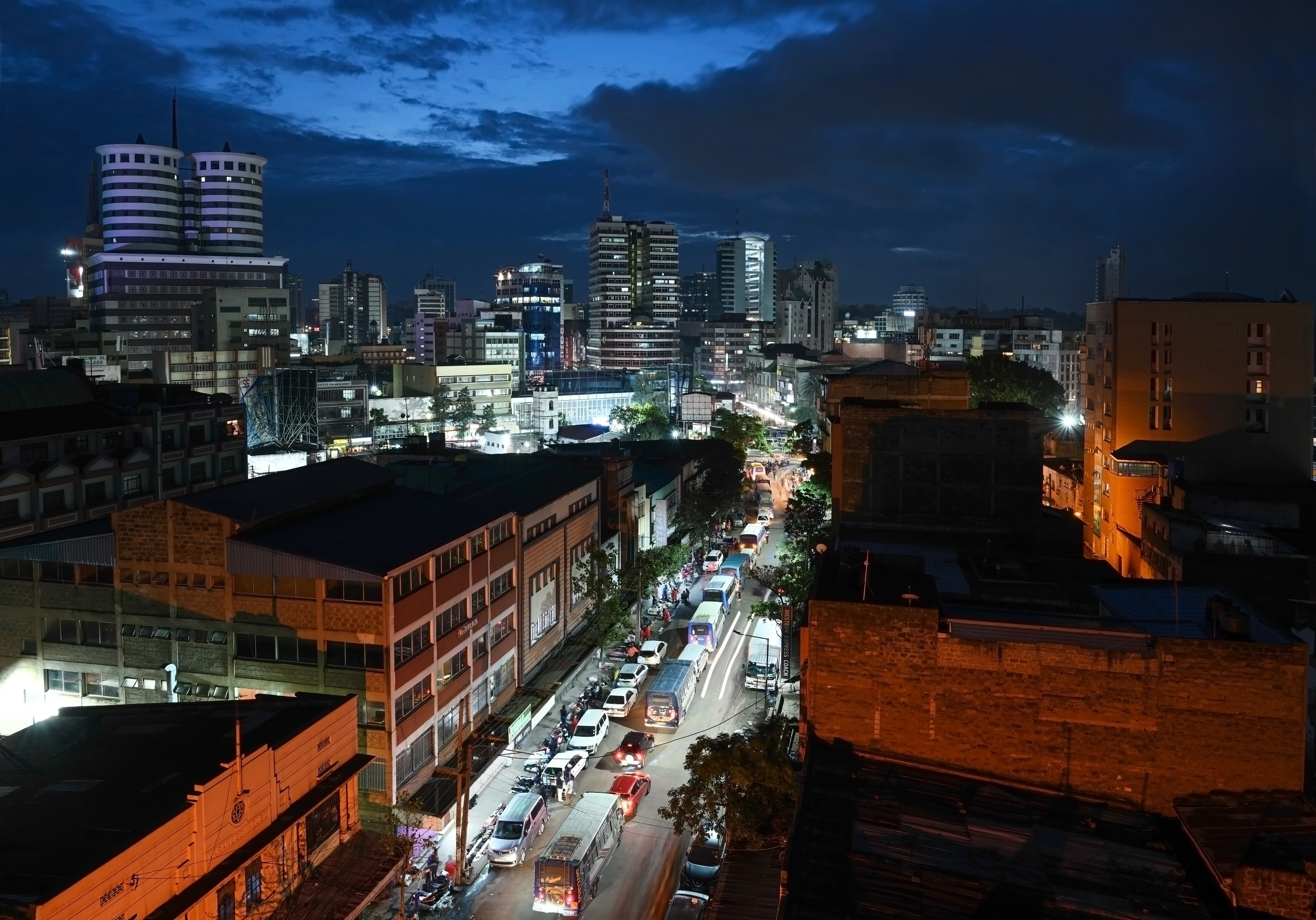 A view looking down from a building of a city street at night, illuminated and filled with cars. The towers of the city skyline are seen in the distance.