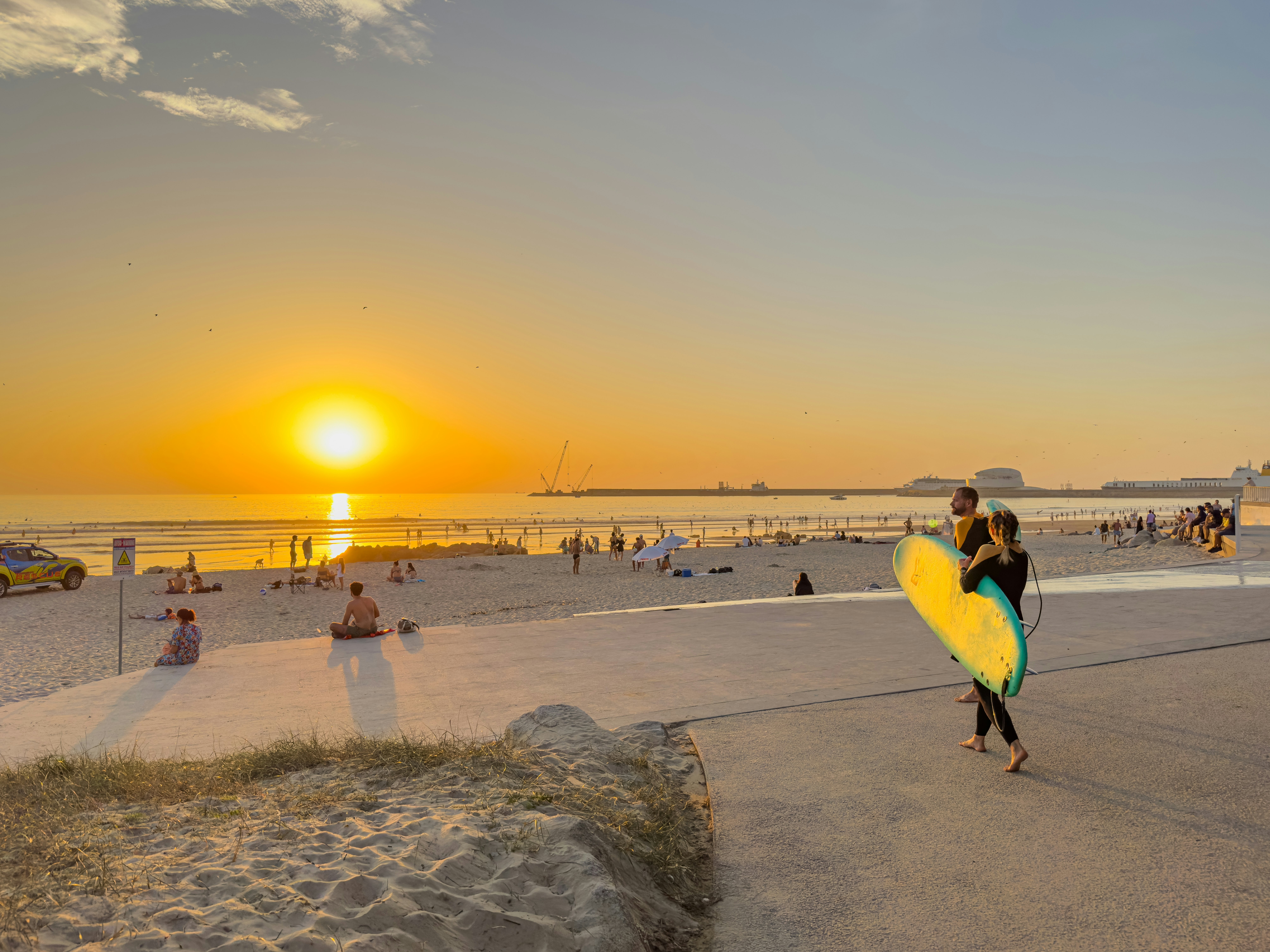 A surfer carries a surfboard toward a beach with people on the sand near Porto, Portugal, at sunset.