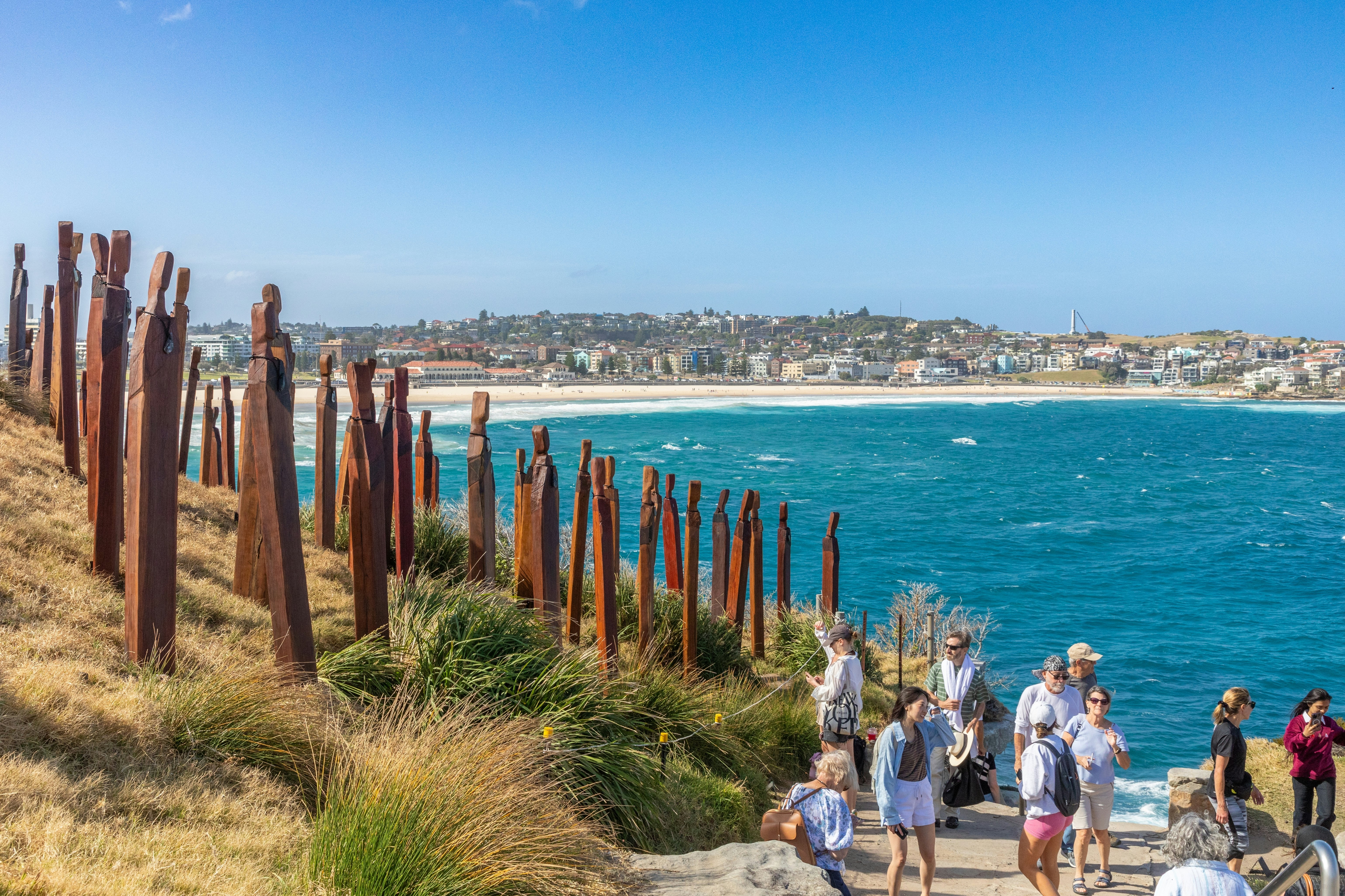 People walk past a sculpture installation shaped like cactuses on a coastal walk near a city beach.