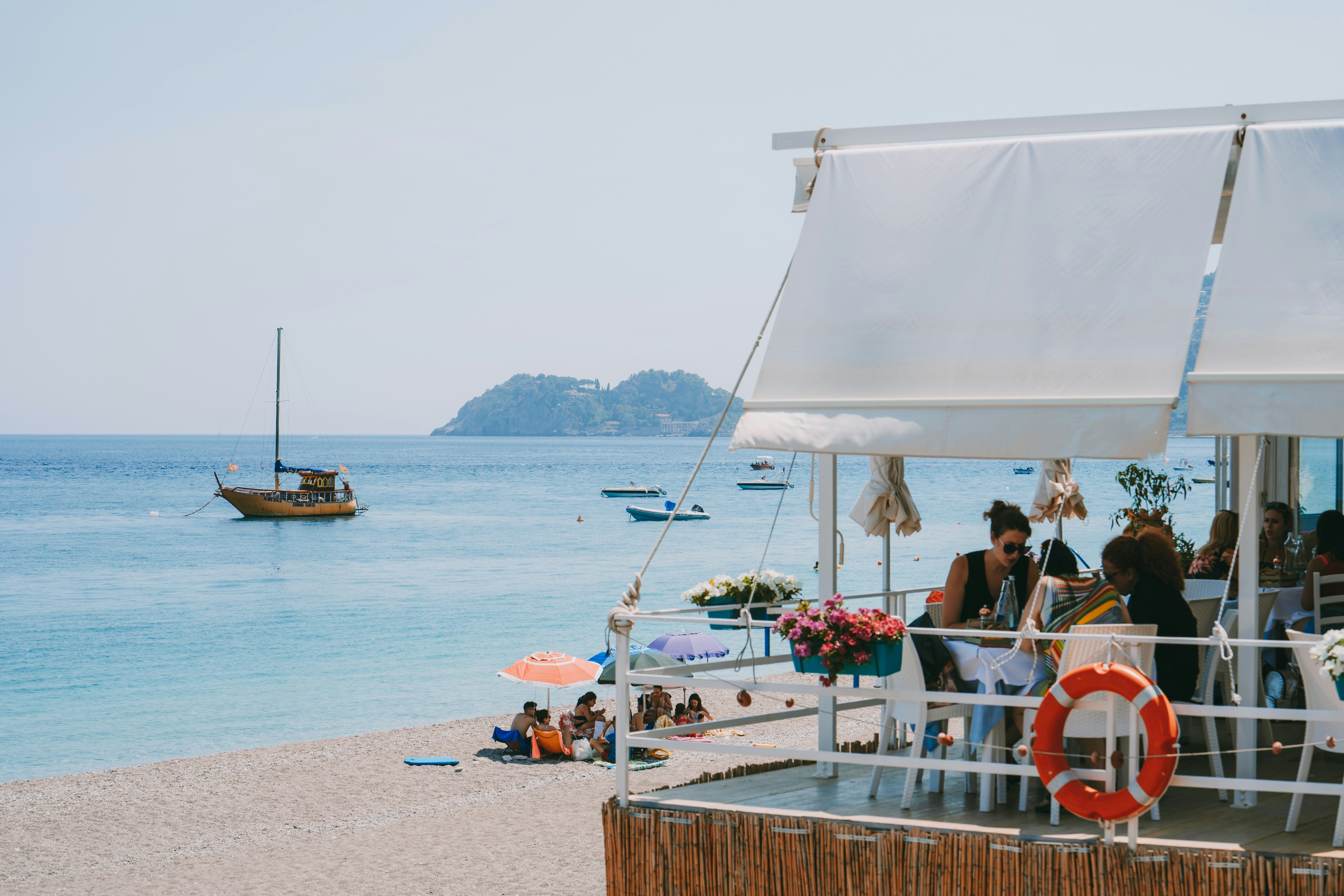 People dine on the terrace of a restaurant overlooking a beach and boats in the water.