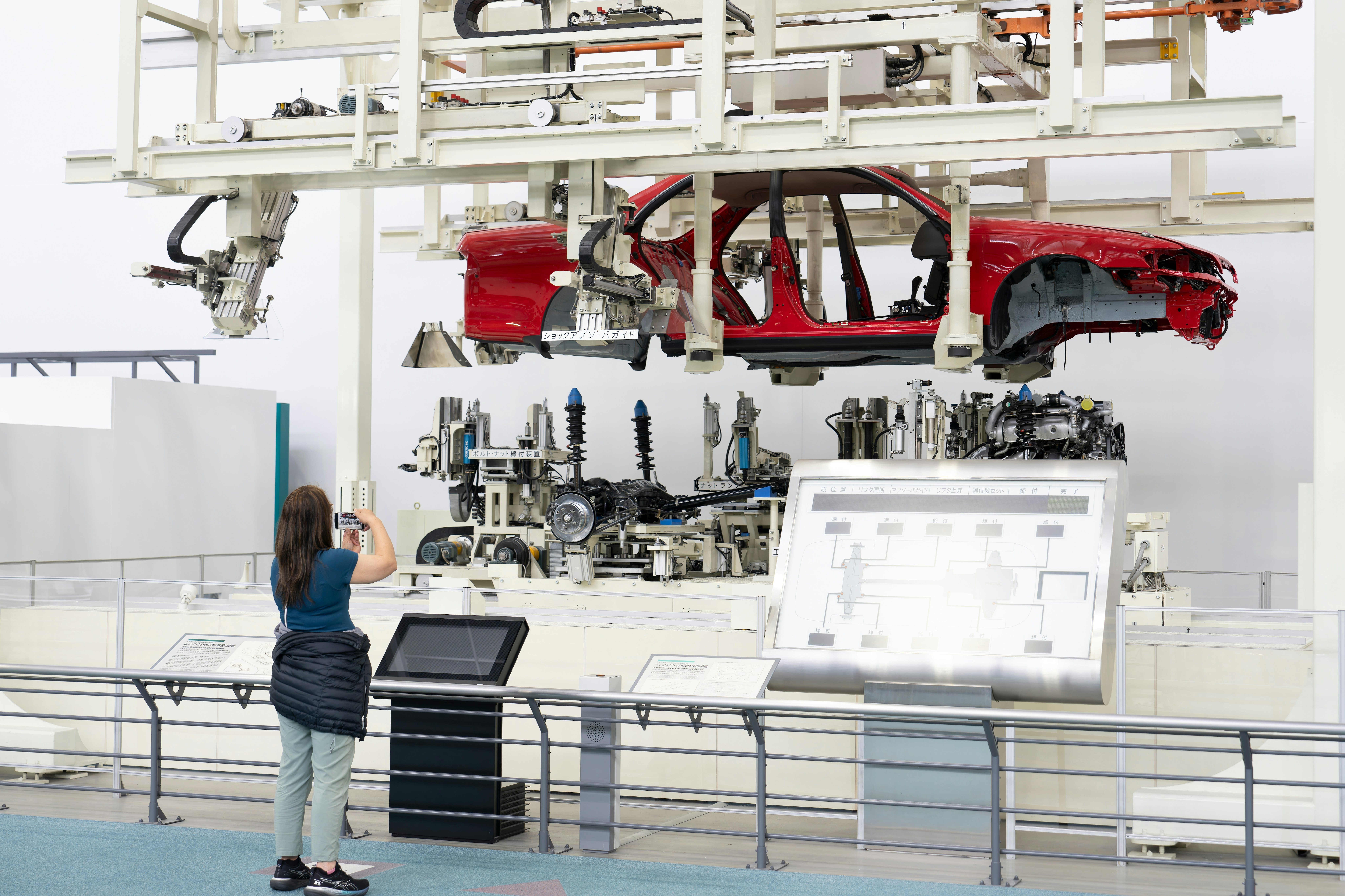 A woman takes a photo of an assembly line with a red car chassis held above the internal parts of the car.