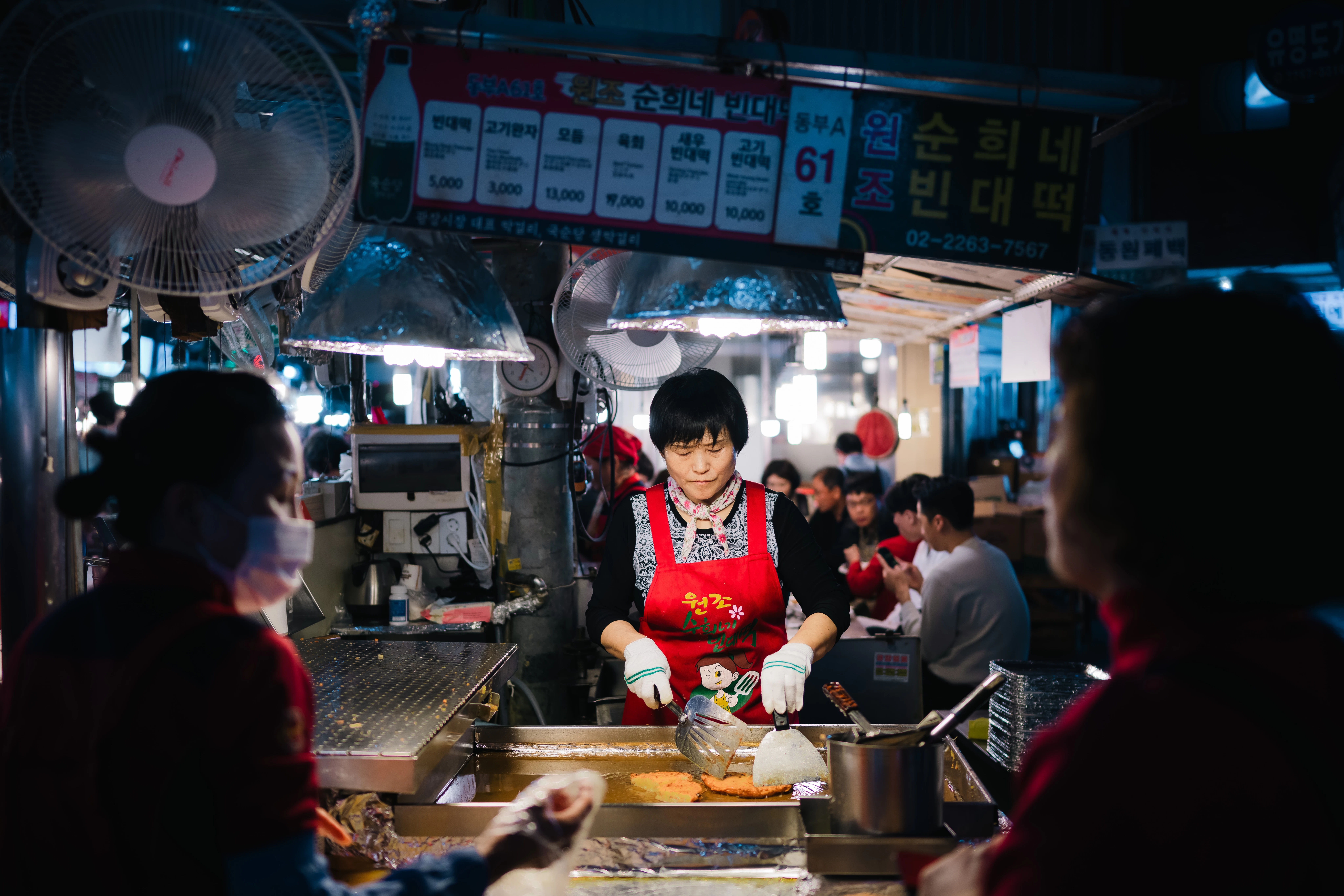 A food vendor prepares meat on a hot plate in a busy food market.