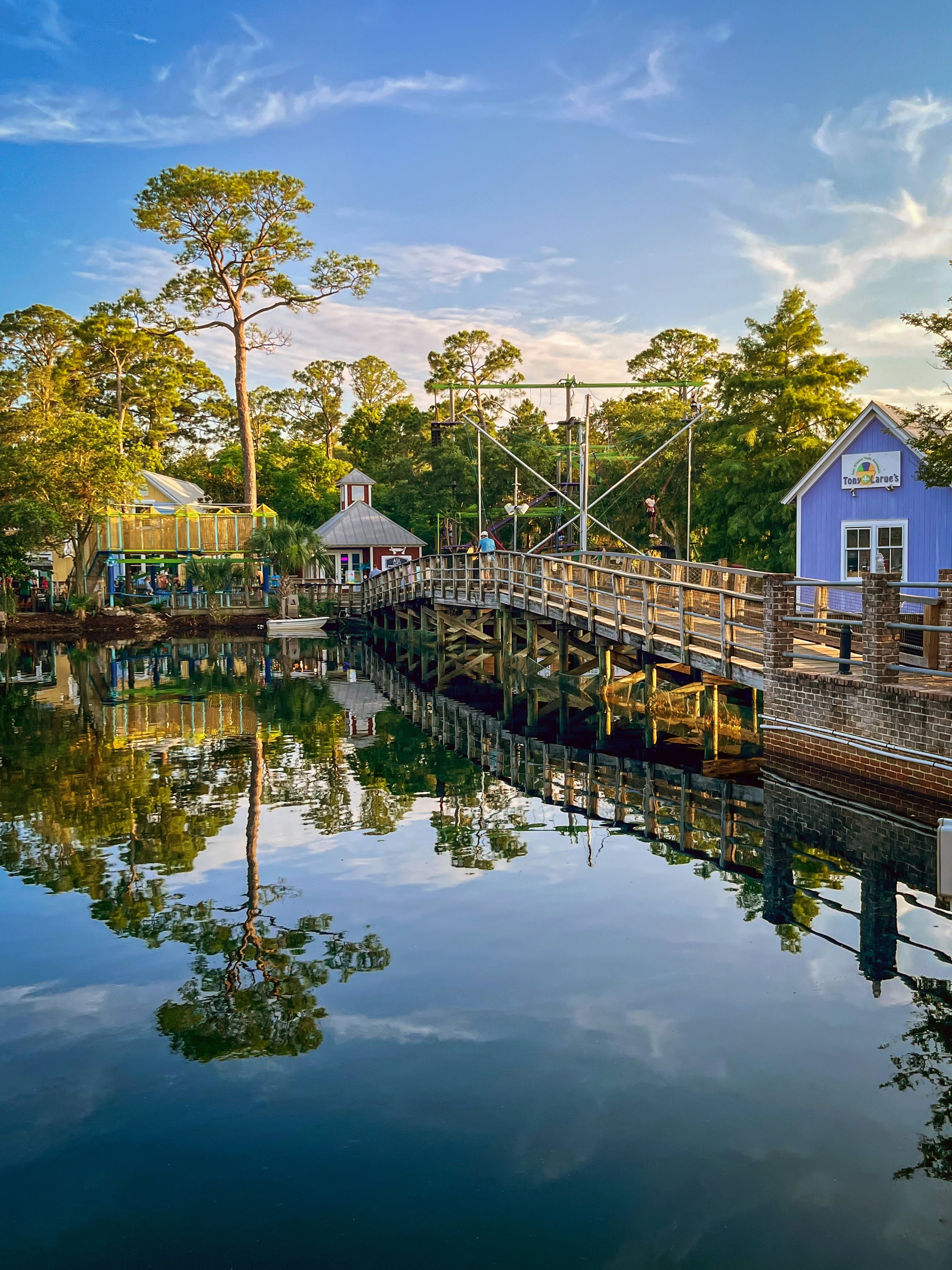 A wooden bridge between two buildings crosses water that reflects the trees and sky.