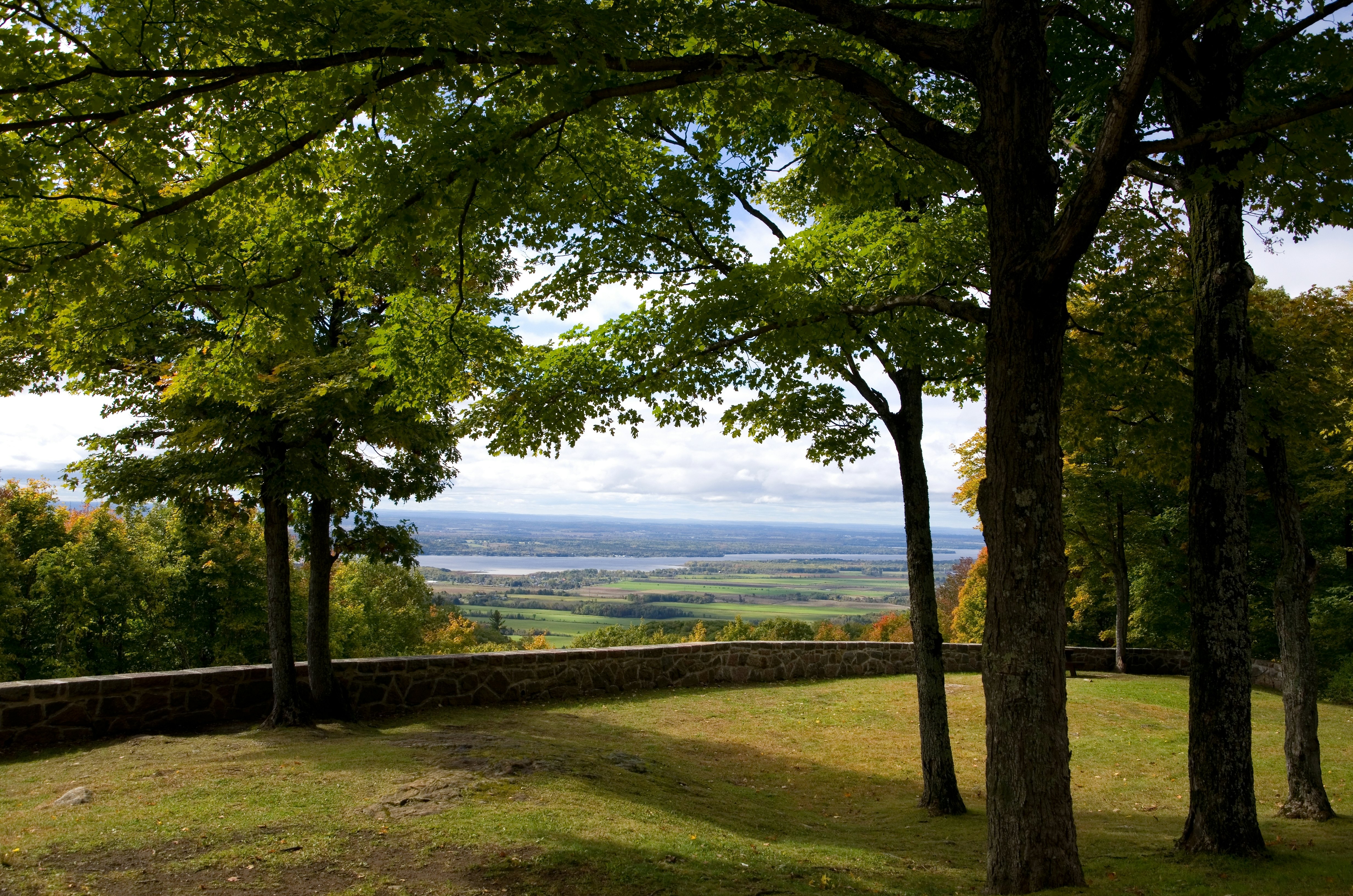 A shaded area in Ontario, Canada, with green grass leads to a low stone wall, beyond which are green fields and a river.