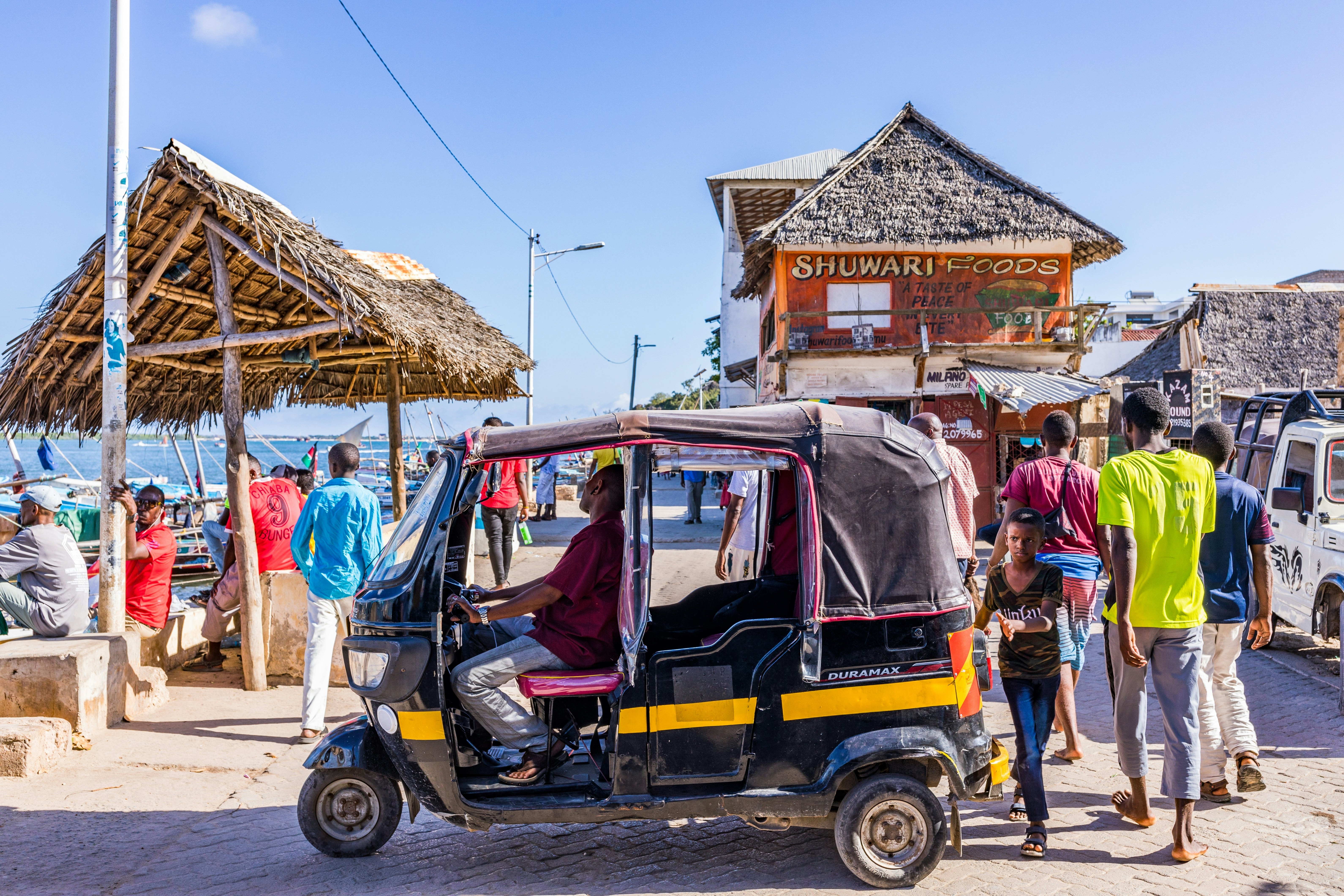 A three-wheeled taxi is parked by a pier in a port, with other people milling around.