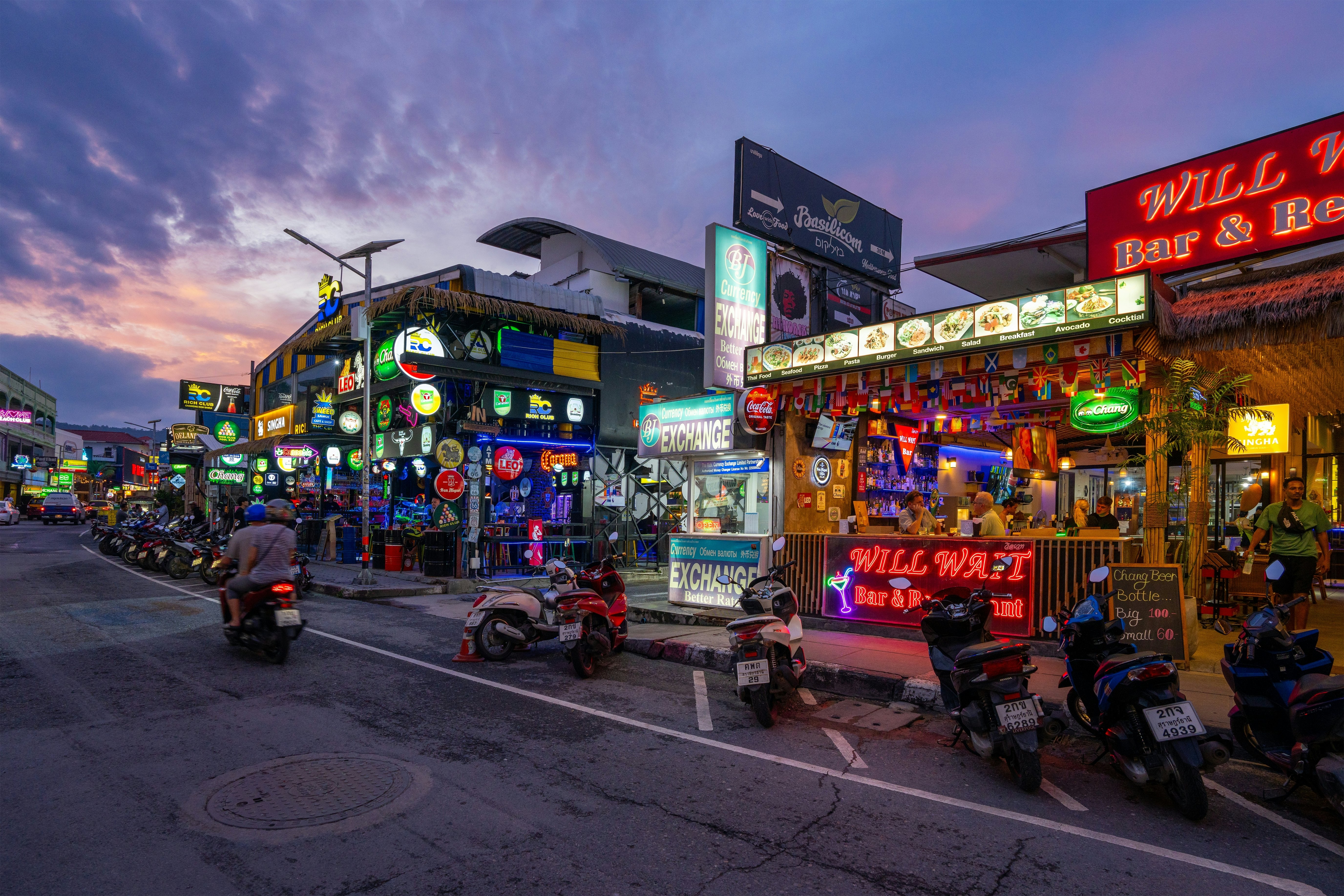 A row of scooters parked in a street lined with restaurants as two motorcycle riders pass by.