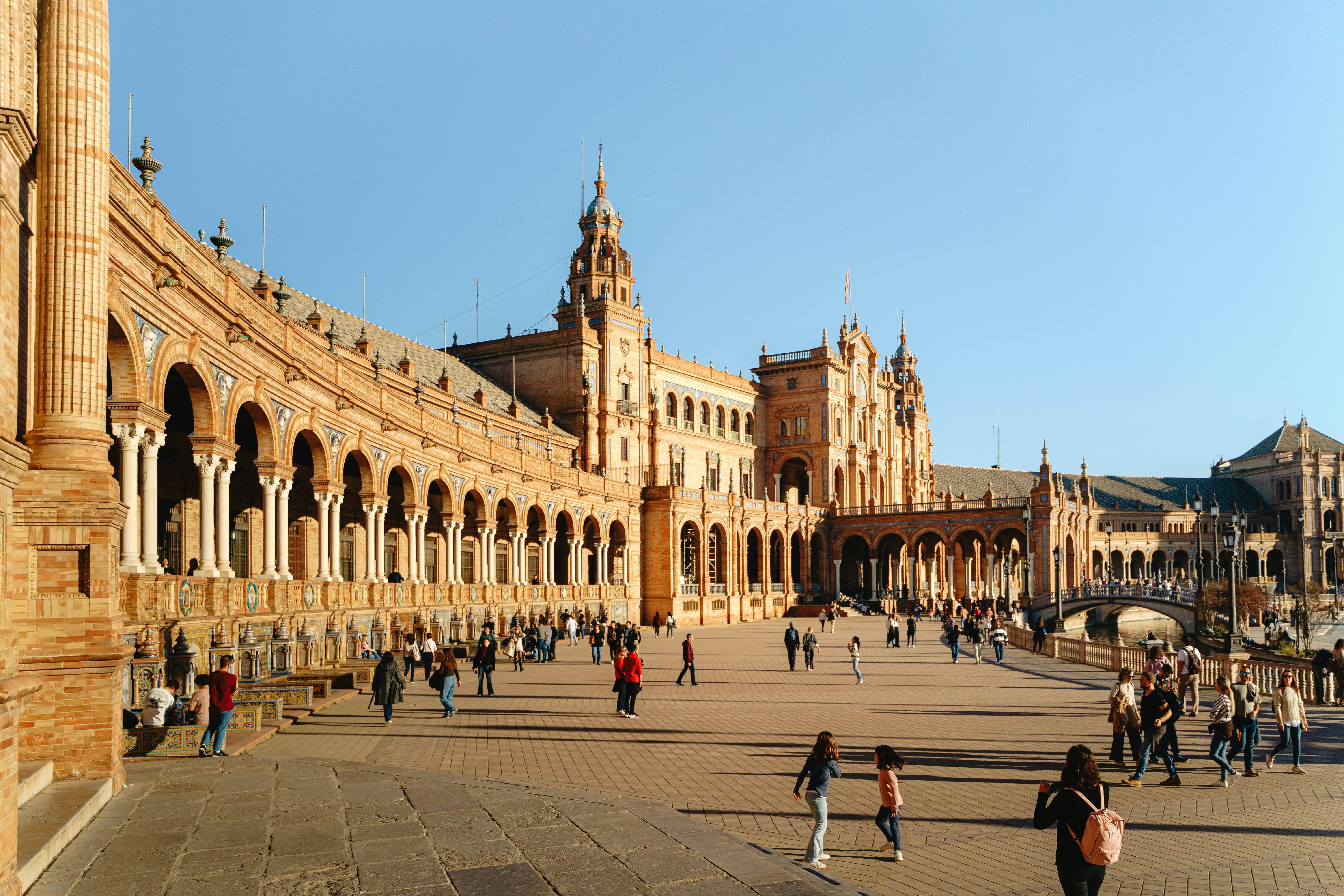 Tourists stroll a semi-circular plaza lined with a mix of Renaissance and Moorish architectural styles, adorned with colorful ceramic tiles.