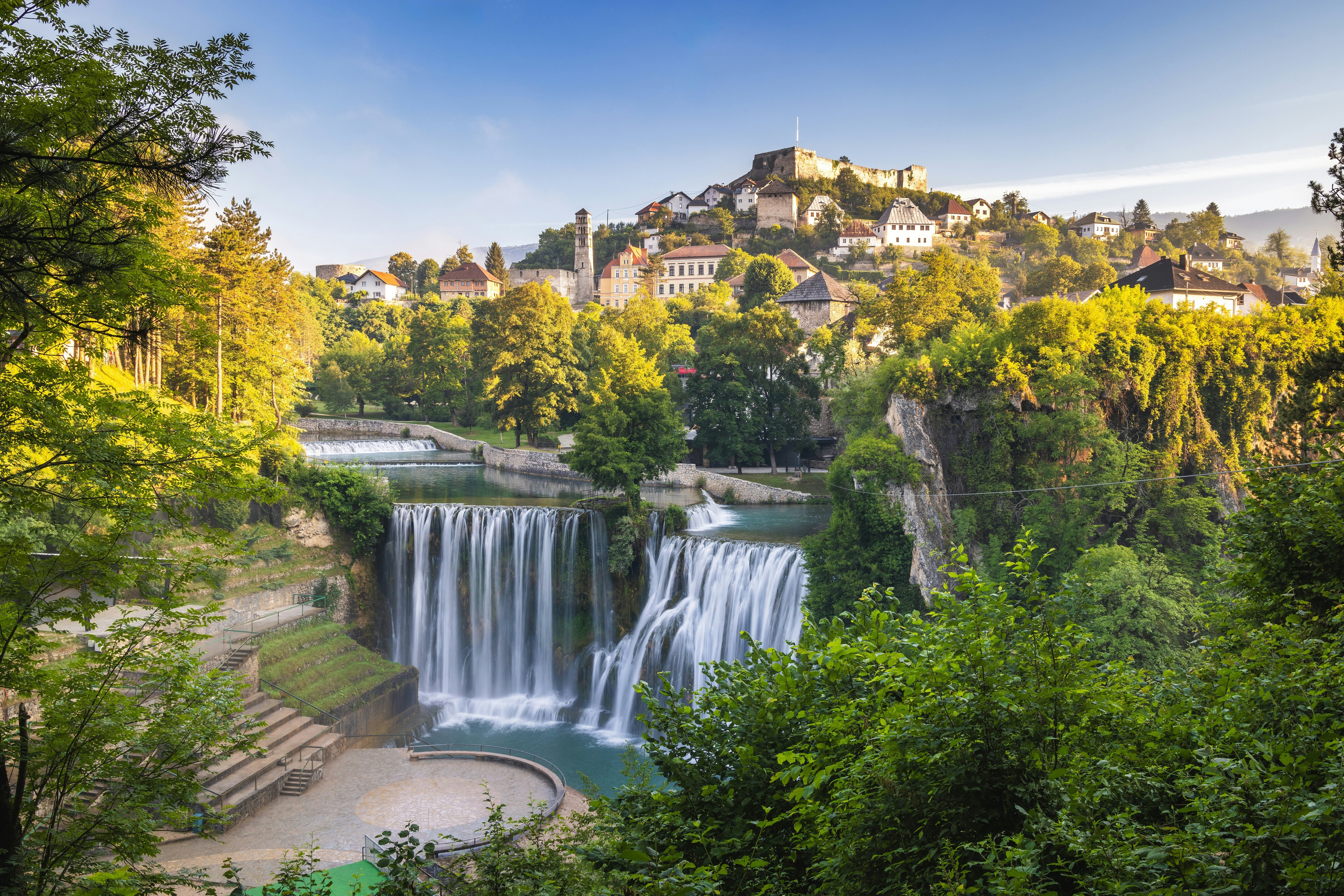 A waterfall cascades into a pool below a hilltop town that glows in the spring sunshine.