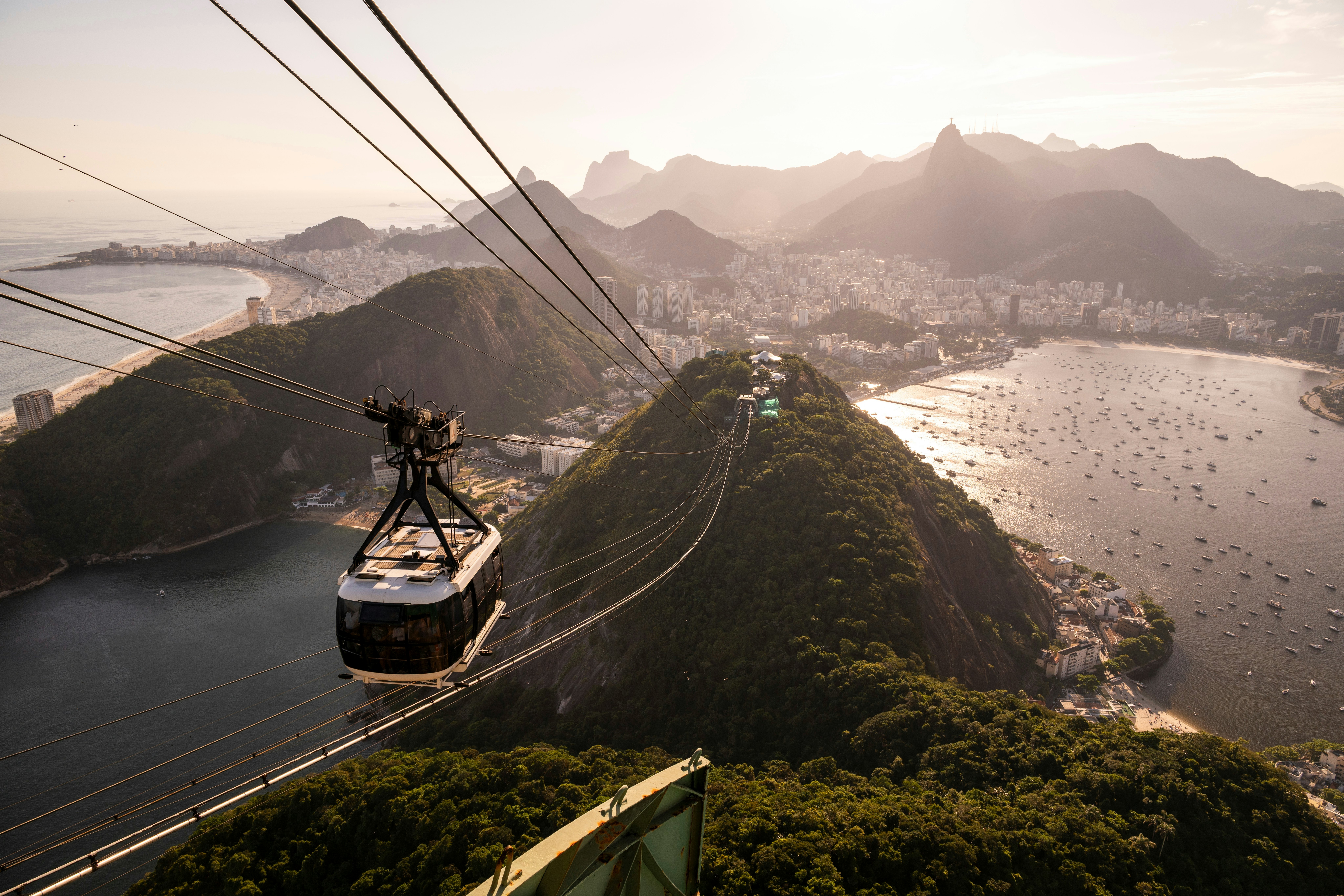 A view from the top of a mountain of looks down on a cable-car gondola and a city beyond, with buildings and beaches in between water and misty mountains.