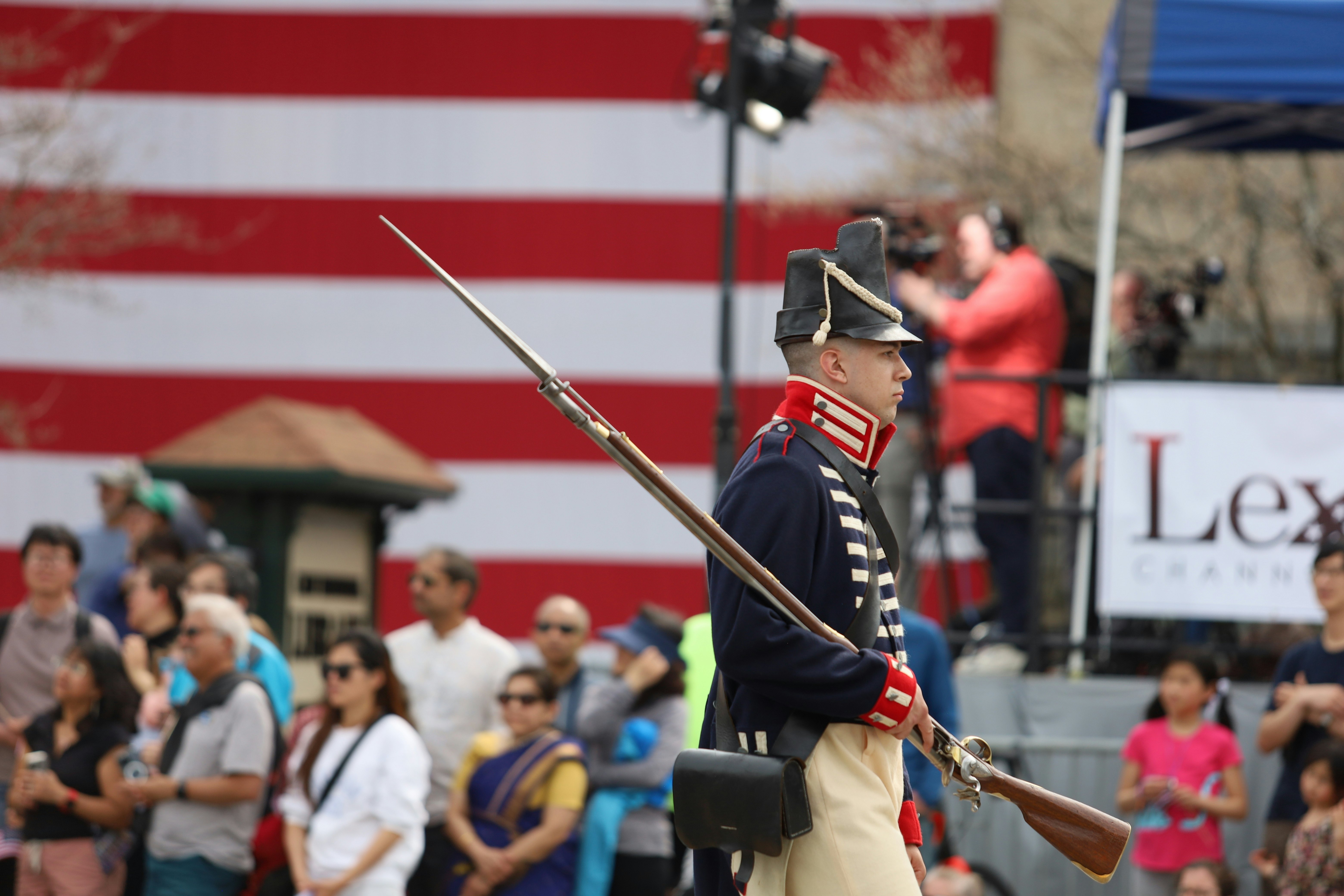 A man in the costume of an 18th-century soldier marches in a commemorative parade in a town street.