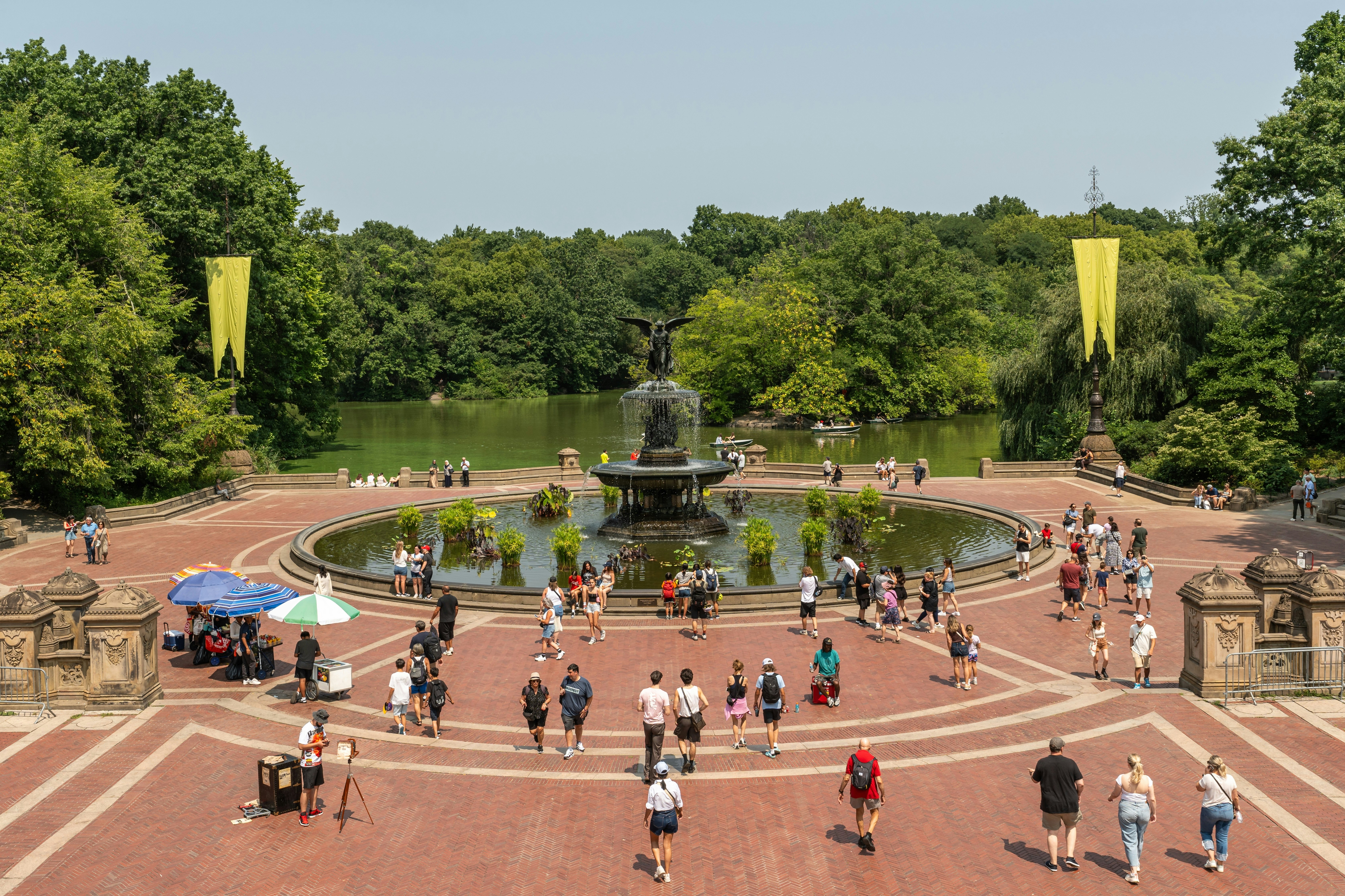 People walking toward and standing around a large round pool of water with a fountain sculpture in the middle at a park under clear daytime skies