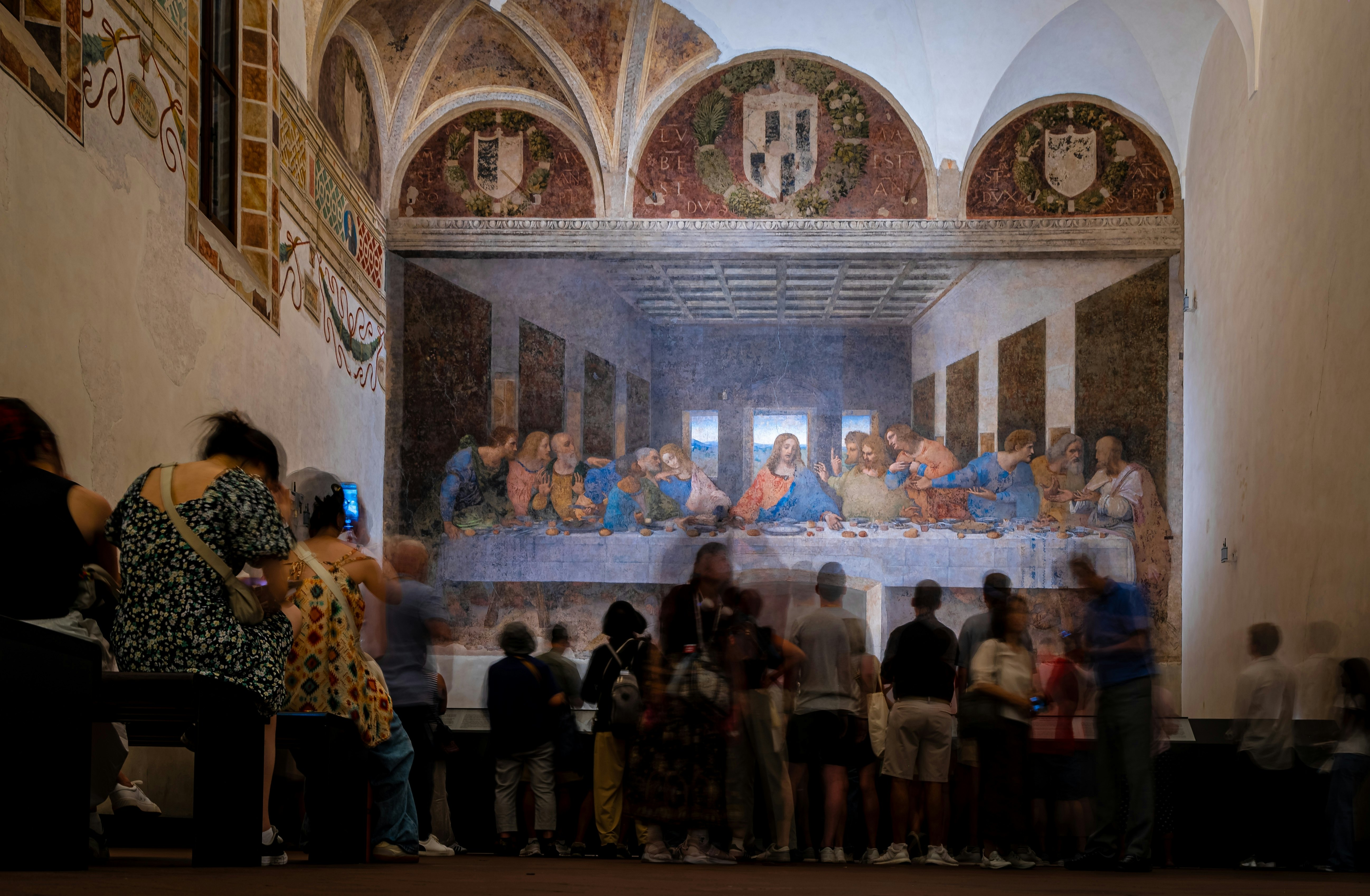 People are seen from behind admiring a fresco covering the wall of a church building.