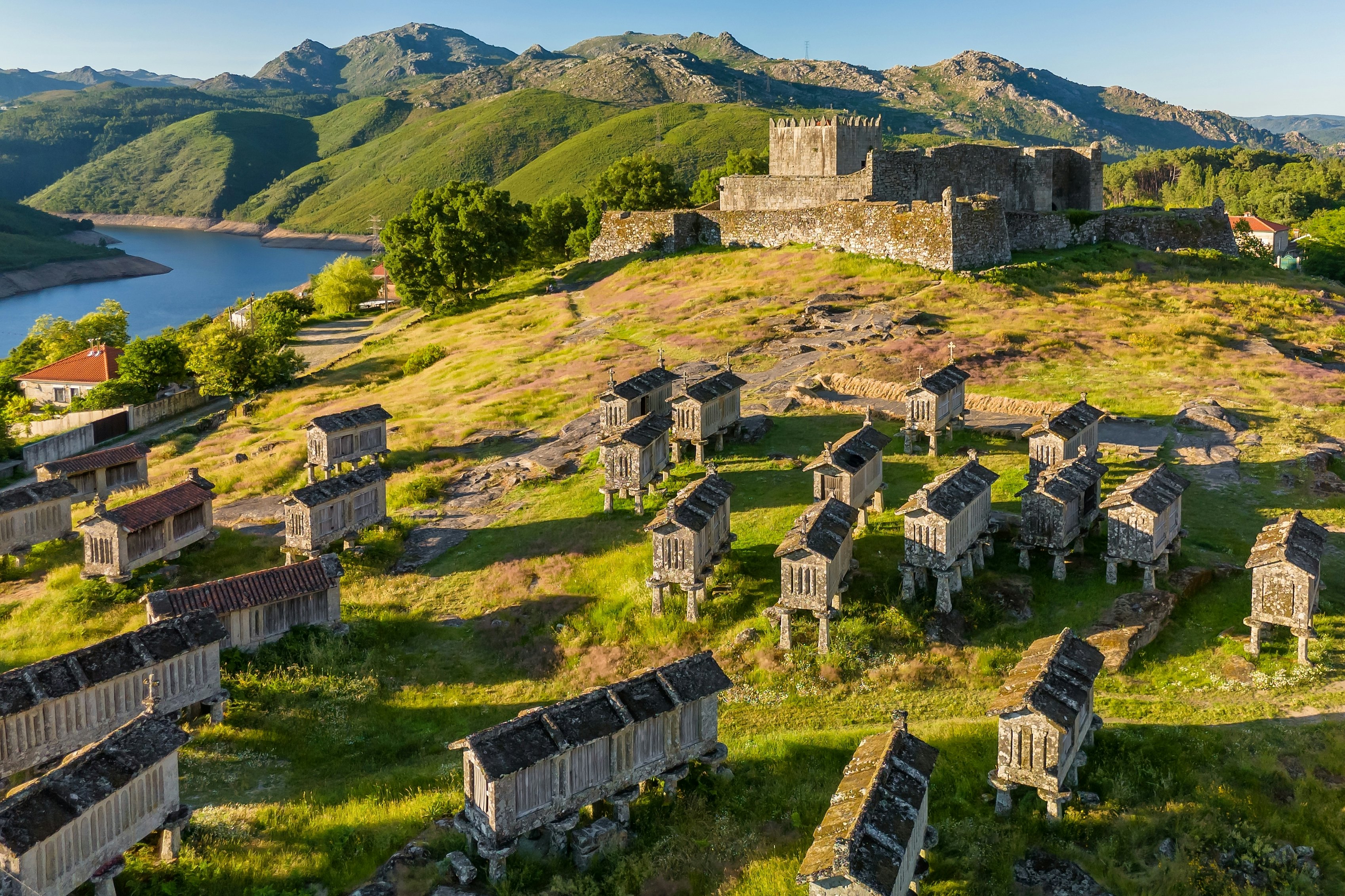 An aerial view of stone granaries in a meadow, with a castle and hills in the background.