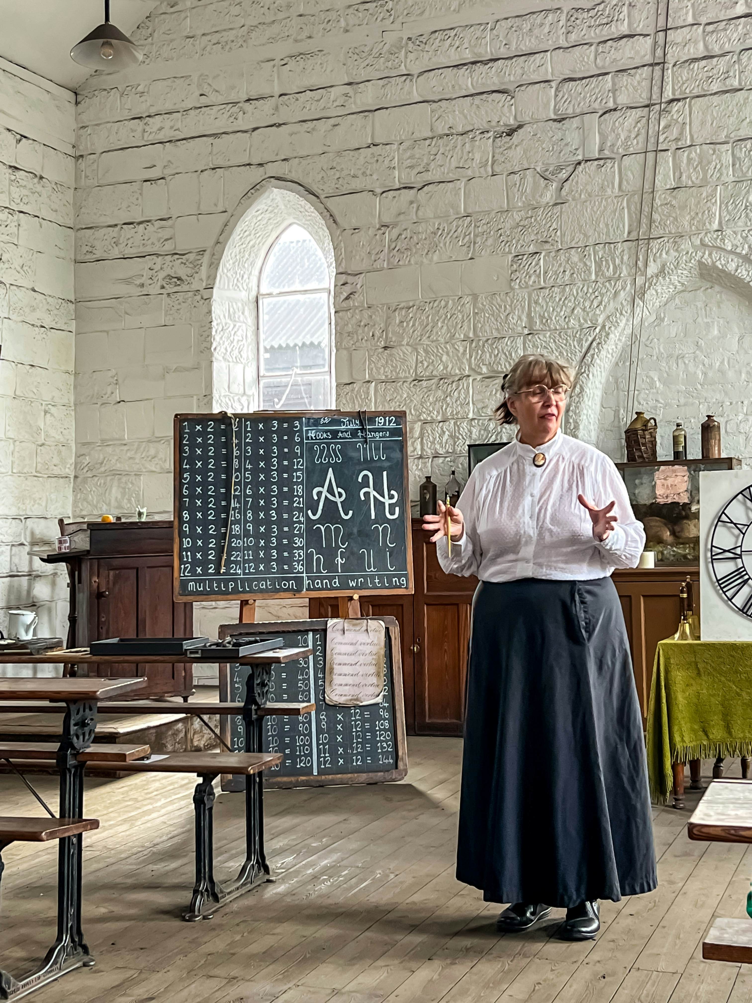 A person dressed in historic clothing stands by a blackboard with a school lesson at a museum in England.