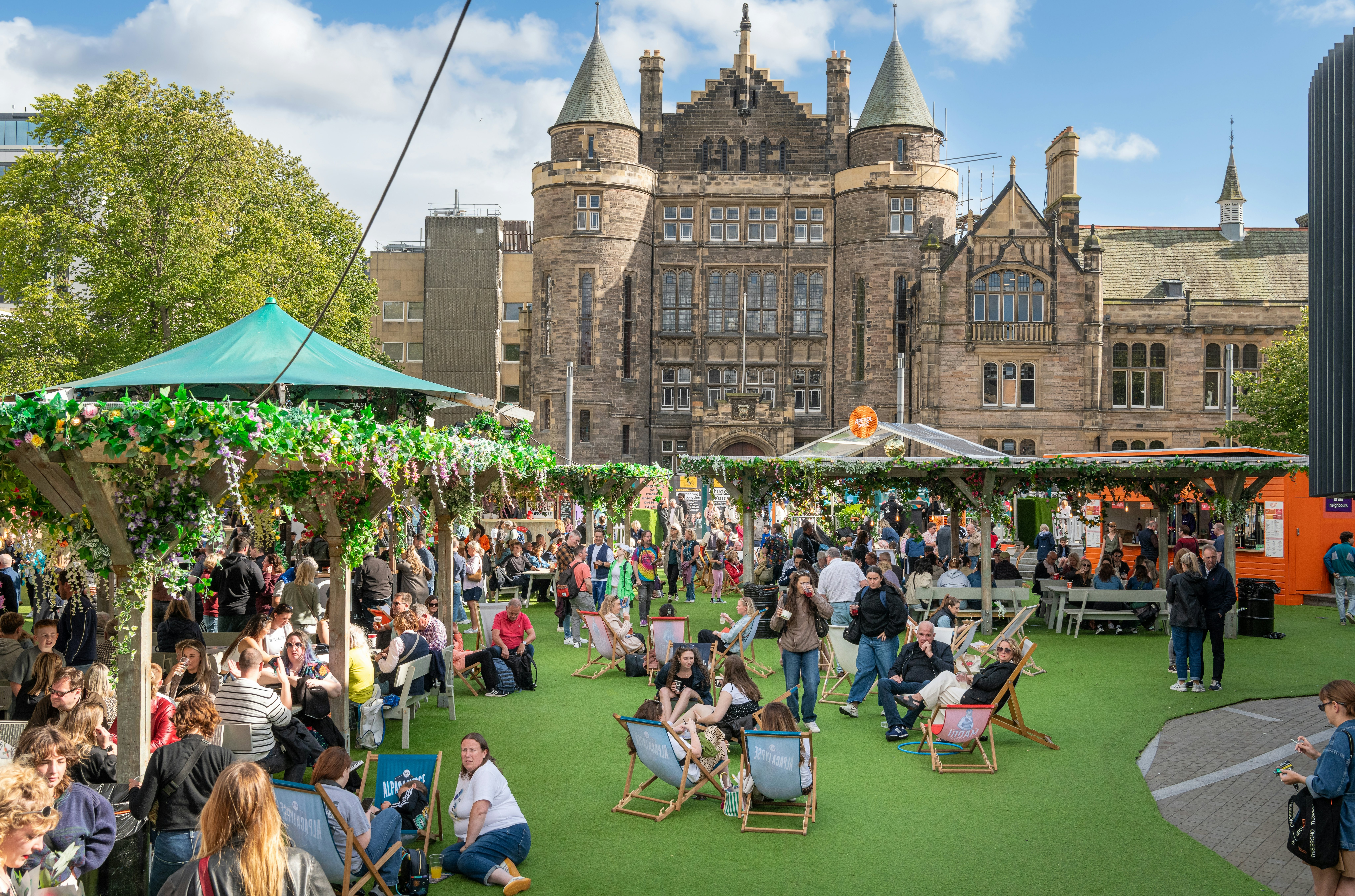 Crowds under arbors and umbrellas in front of a historic building in Edinburgh during the Fringe festival.