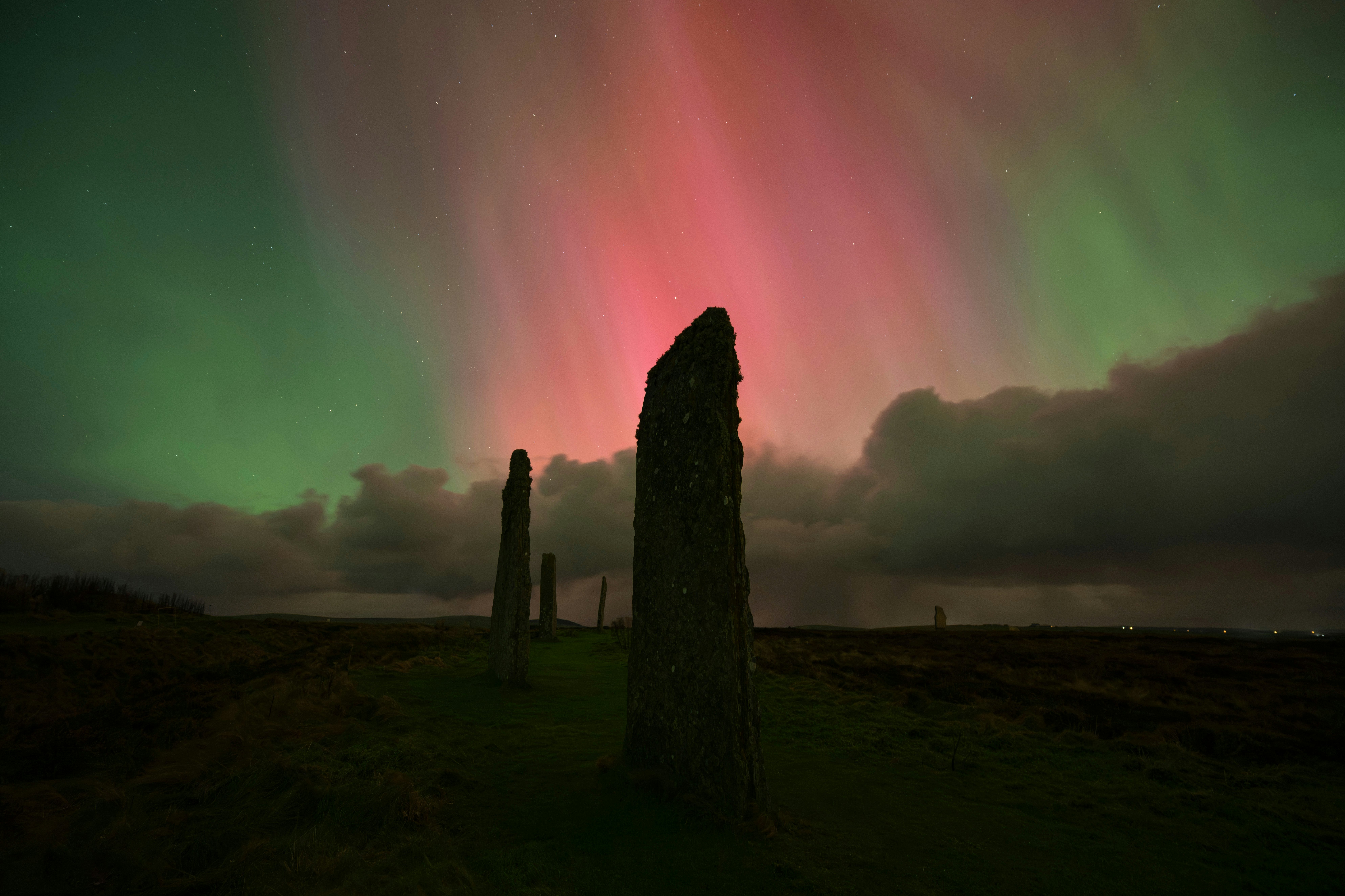 Ancient standing stones on a dark landscape with pink and green northern lights against clouds overhead.
