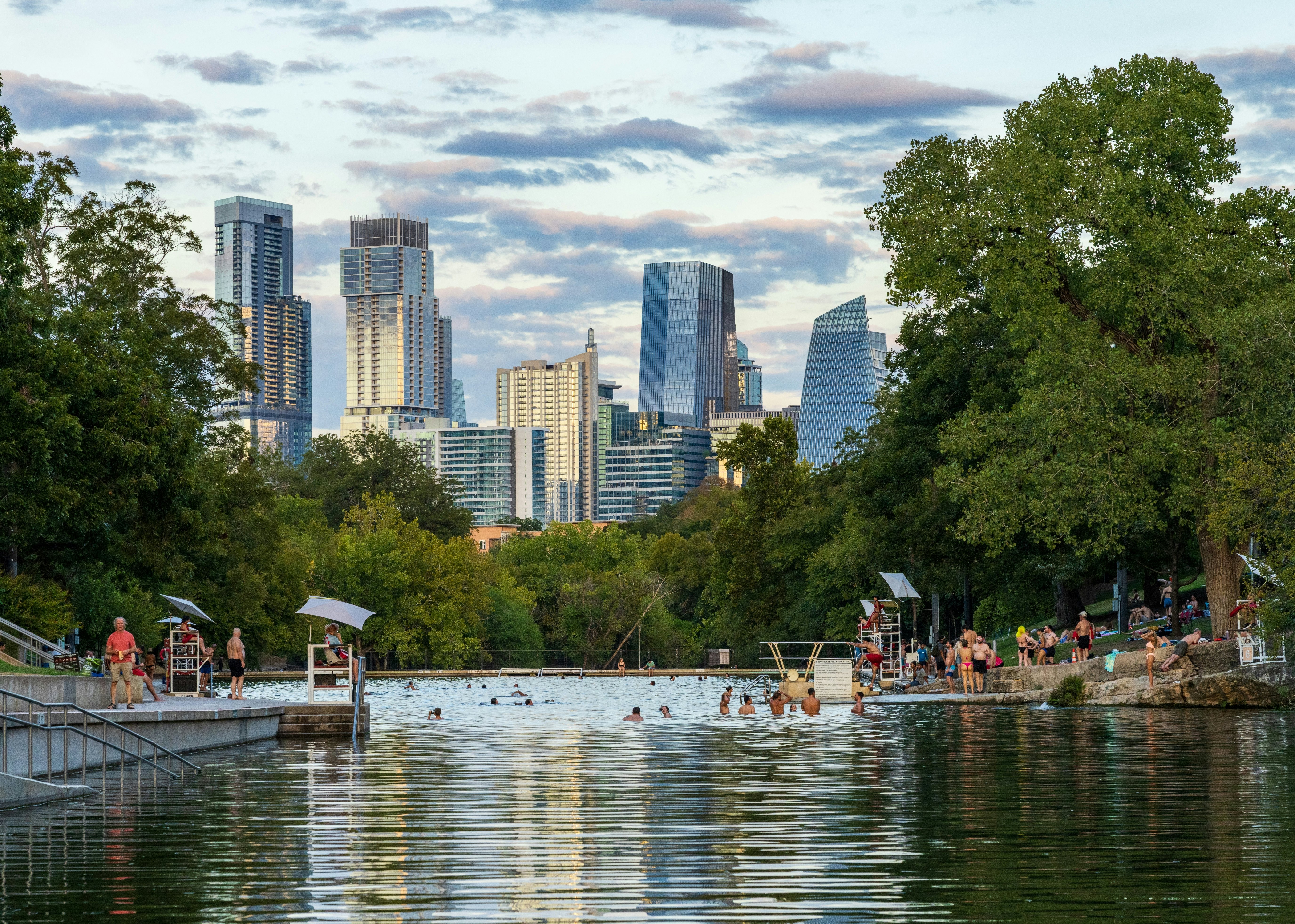 People swim in a natural pool shaded by trees, with the skyscrapers of a city visible in the background.