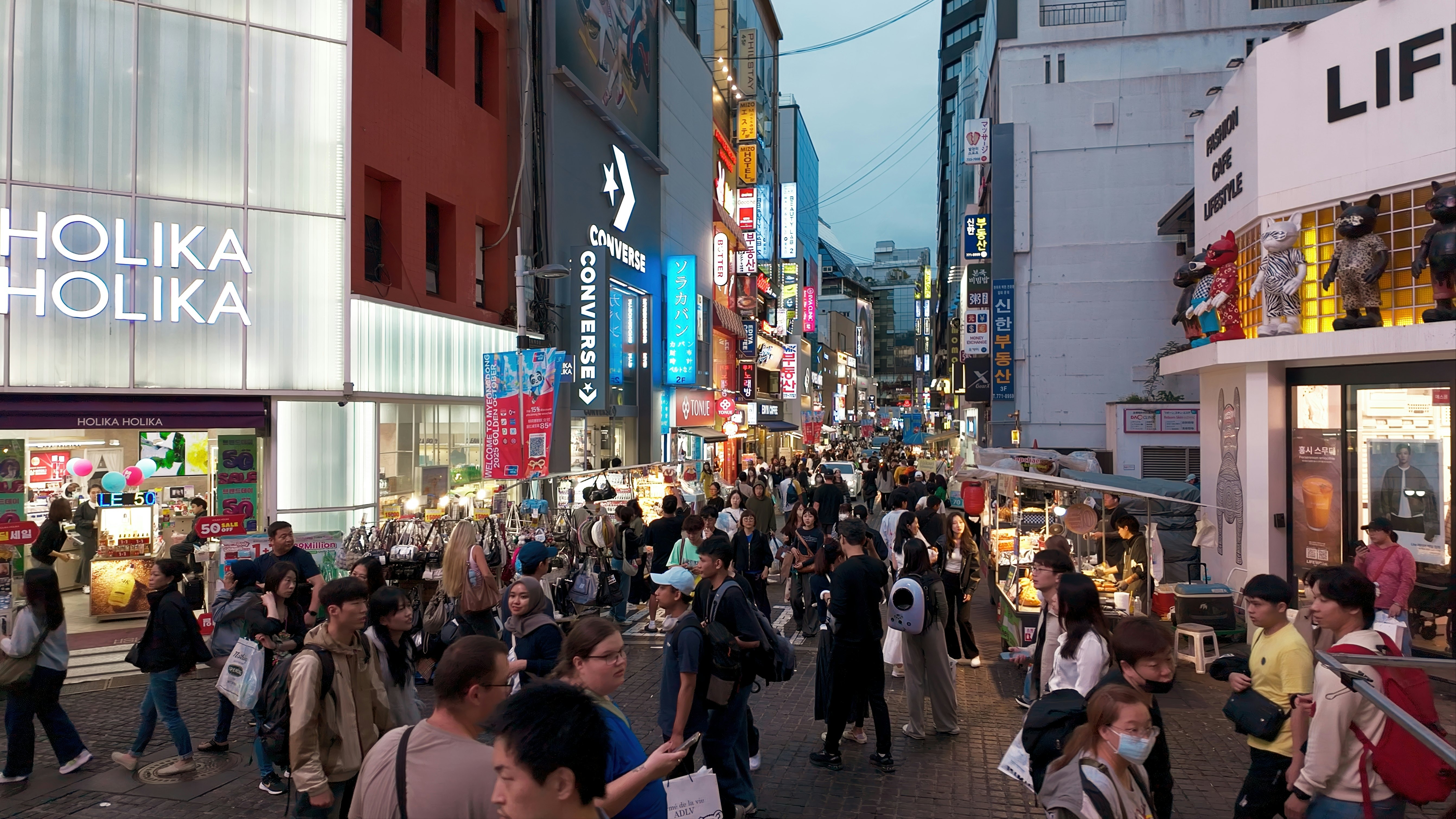Crowds of people walk in a pedestrianized street at dusk. The lights of stores and signs illuminate the scene.