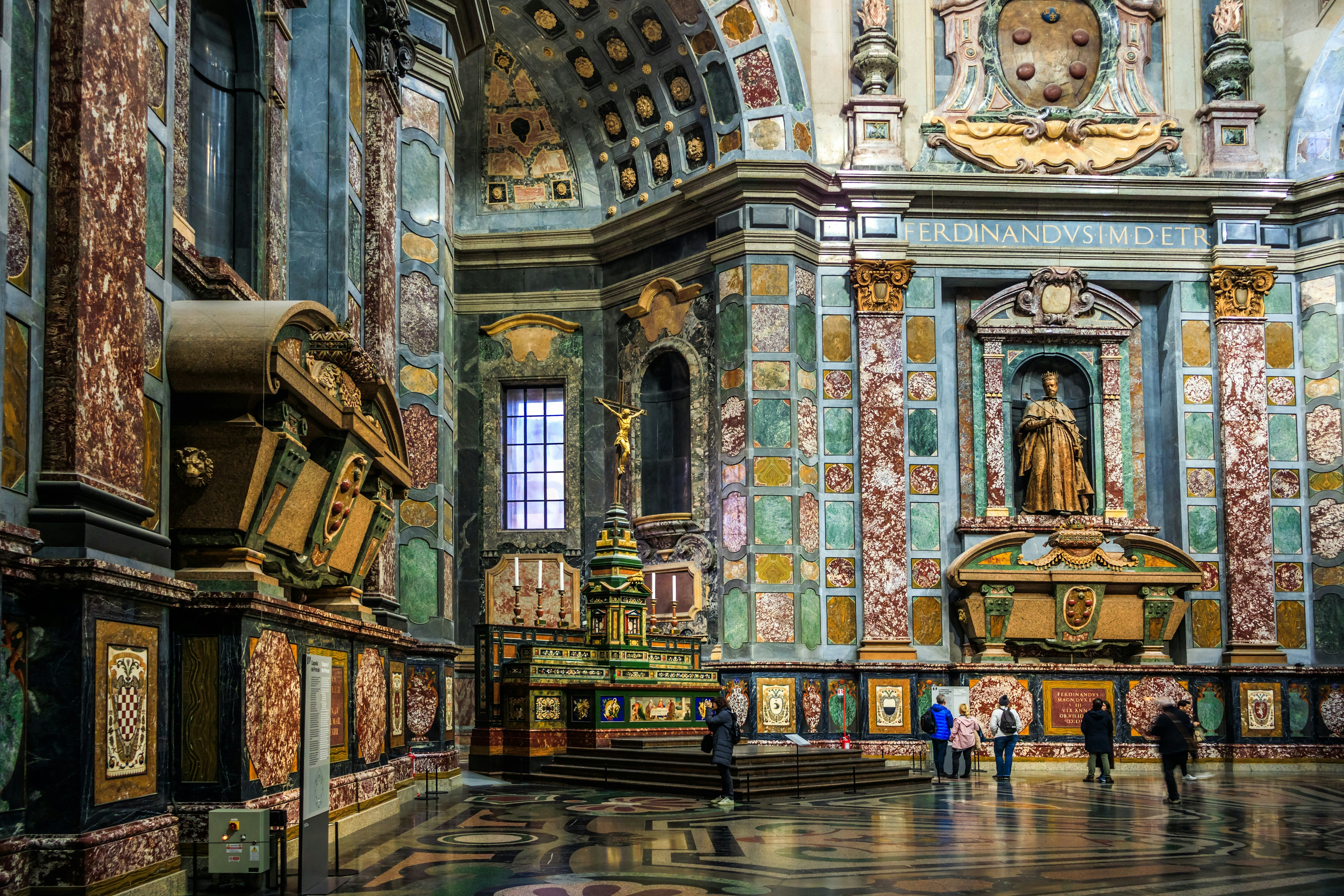 People stand in a huge chapel, its walls covered with polychrome marble and lined with elaborate tombs.
