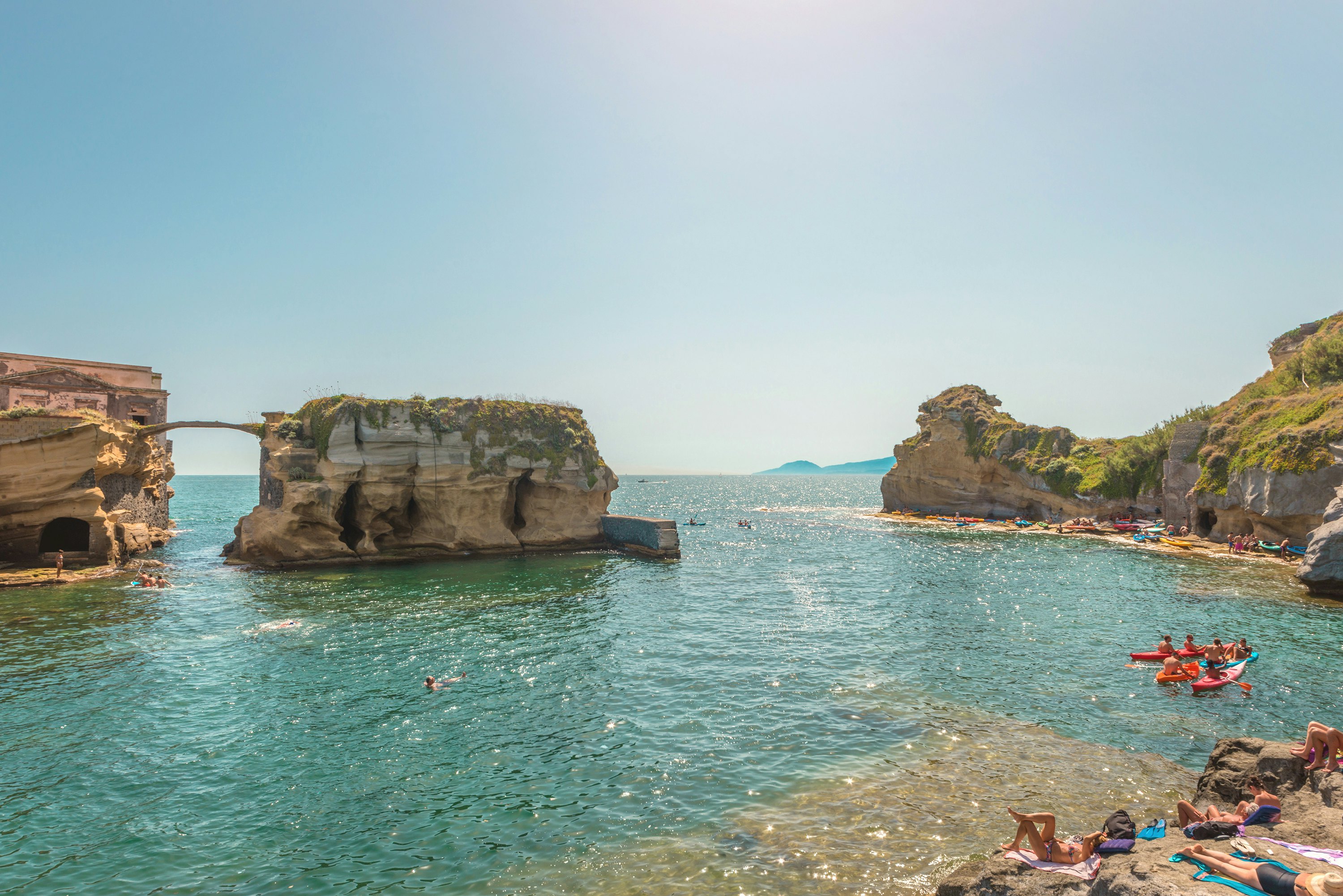 A wide view of a shallow cove, with rock islets in the water, and kayakers seen by the shore to the right.