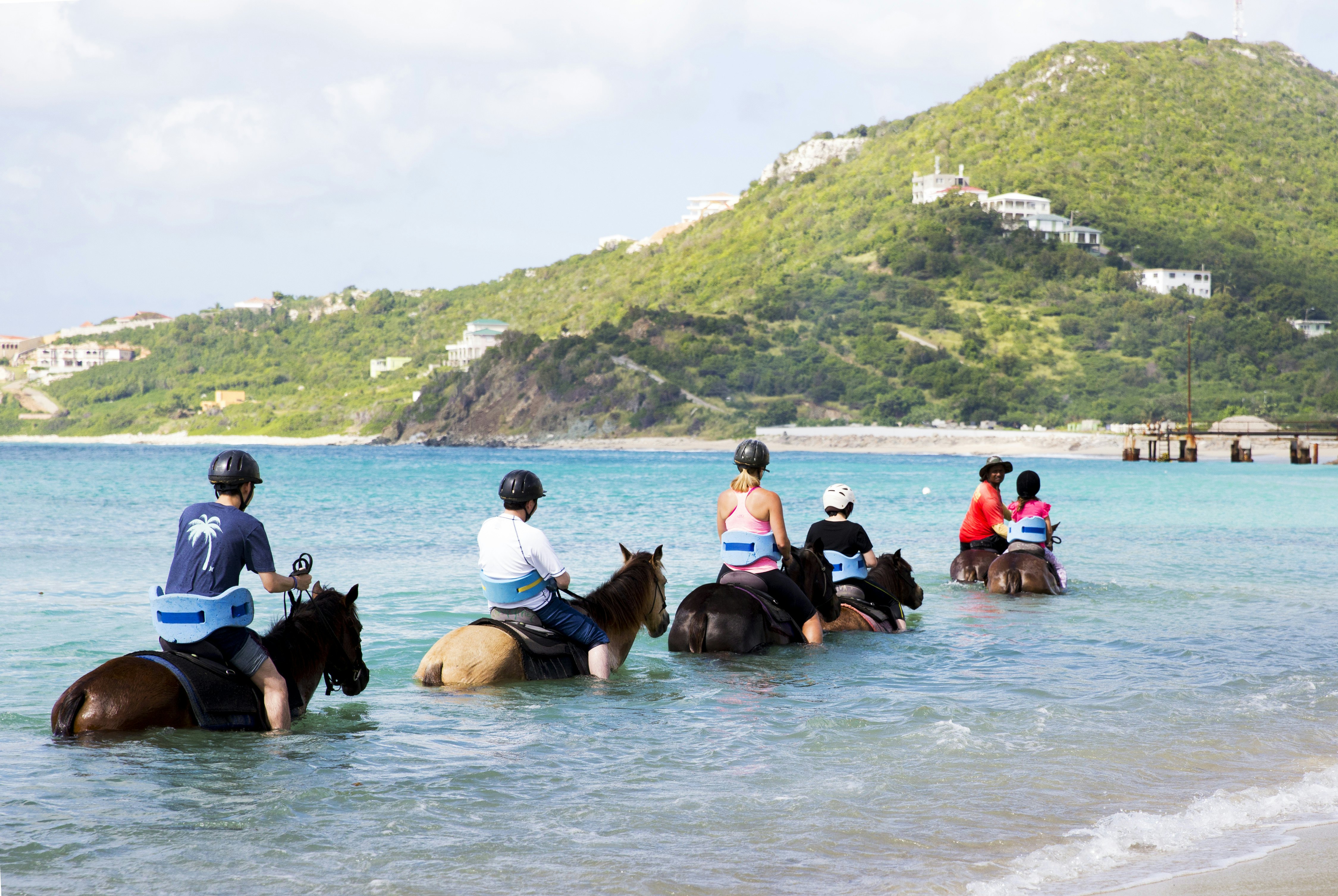 A group of riders on semi-submerged horses in the surf off a tropical beach.