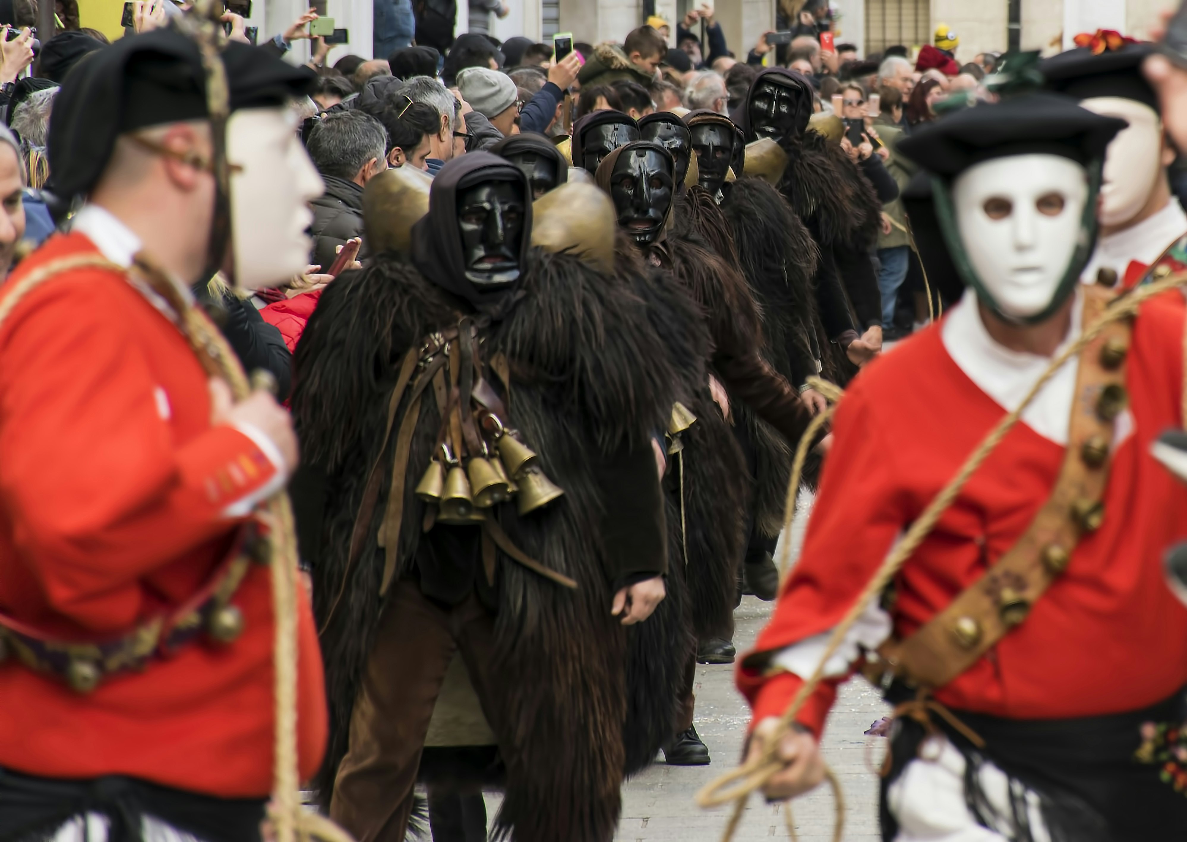 Men wearing black face masks and dressed in fur with bells round their neck parade down a street, with others in red jackets and white face masks standing nearby.