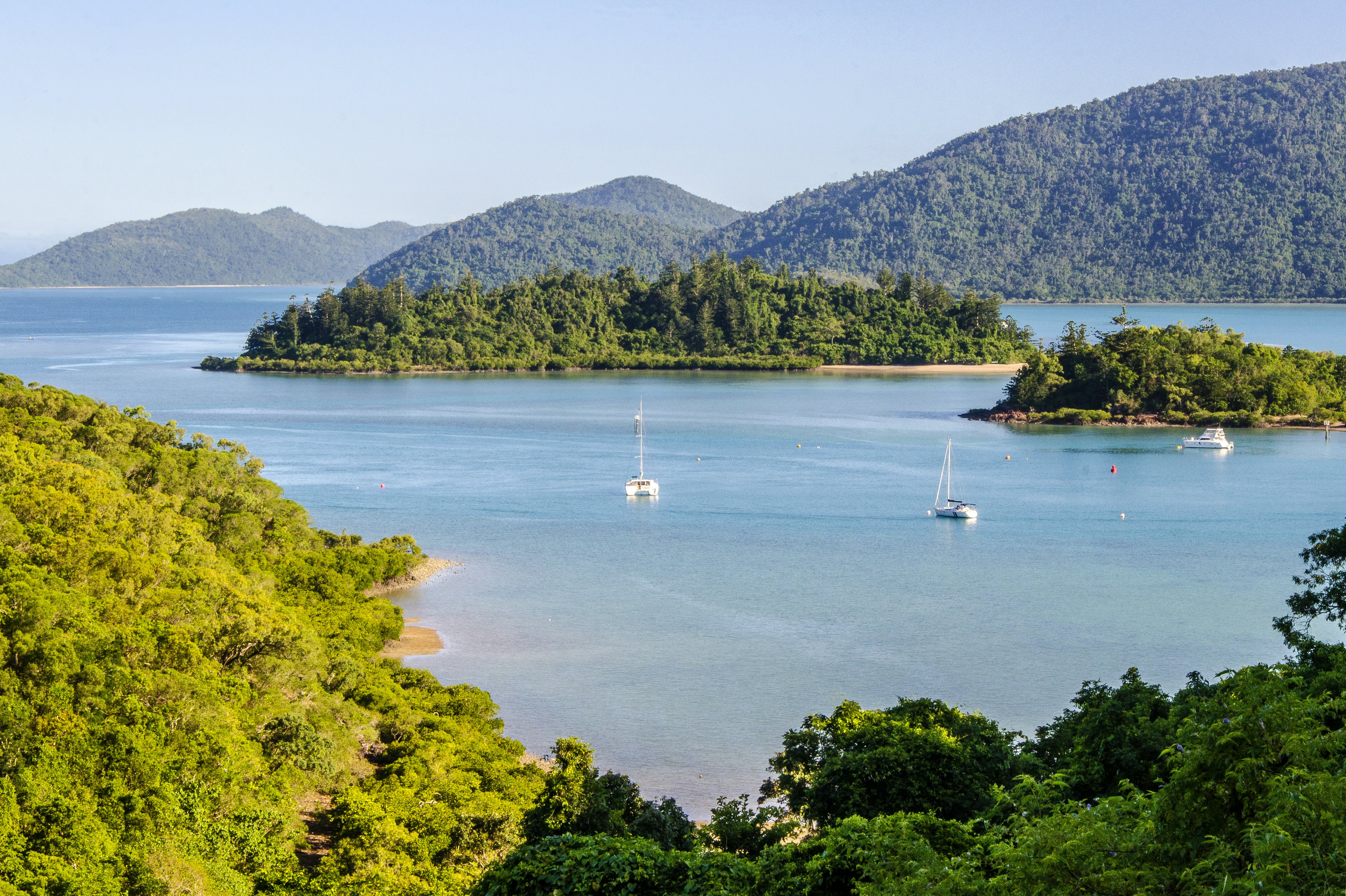 Boats in a blue cove, with green islands in the water and forested hills in the foreground and background.