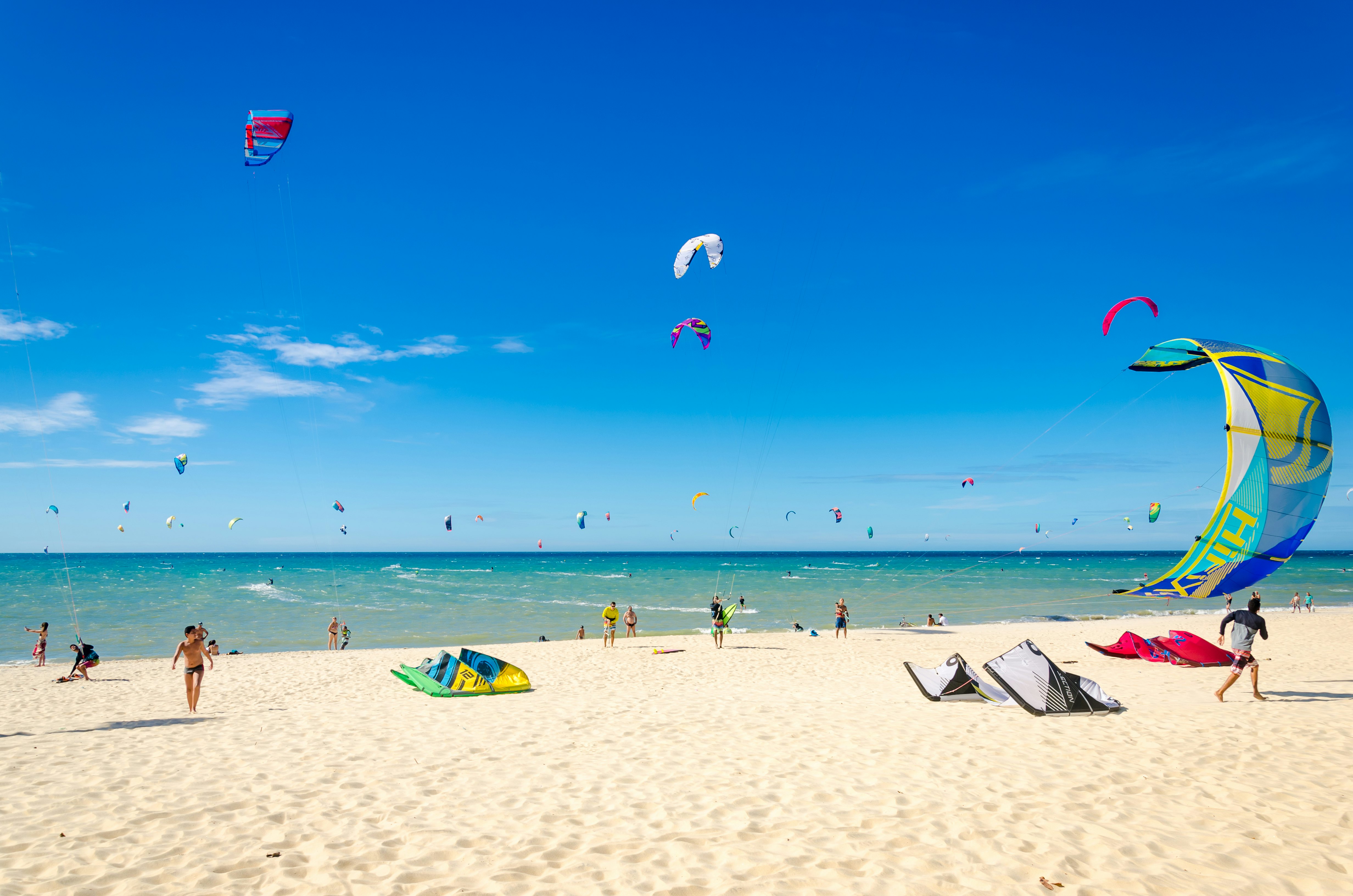 A white-sand beach with people setting up colorful canopies for kitesurfing.