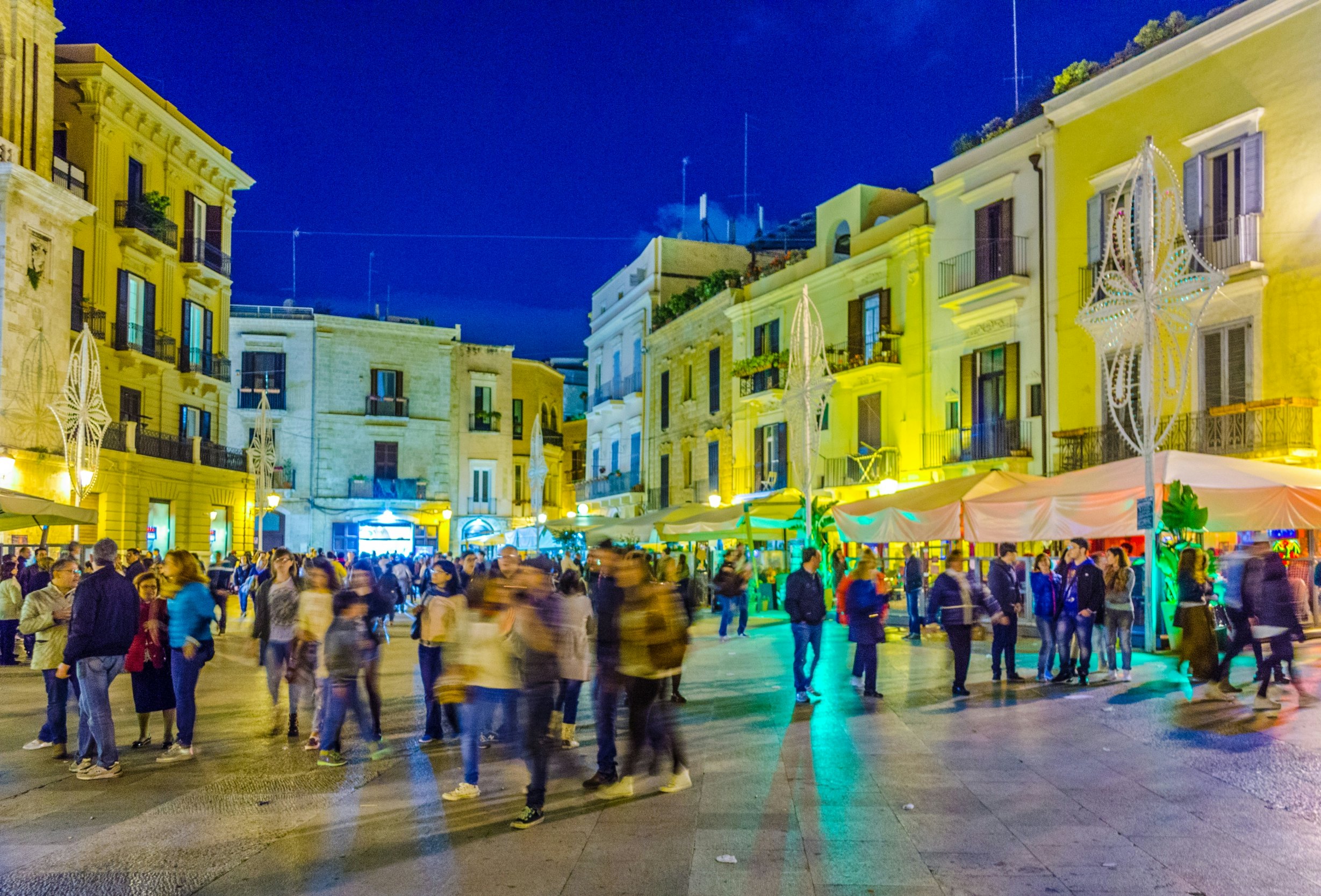 People mill in a city square at dusk. Lights from the buildings and restaurants provide a colorful glow.