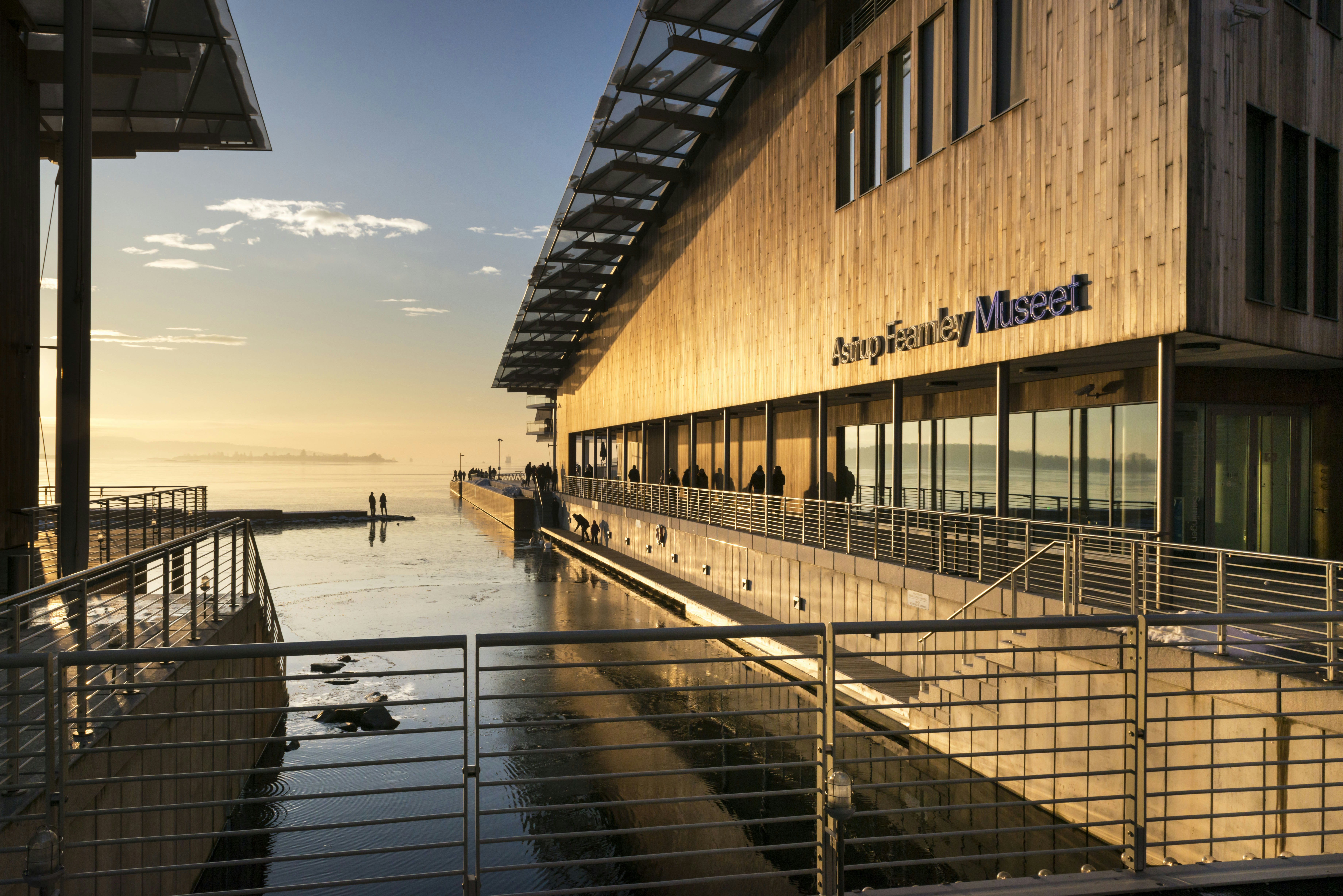 A harborside museum building with people strolling along the walkway at sunset.