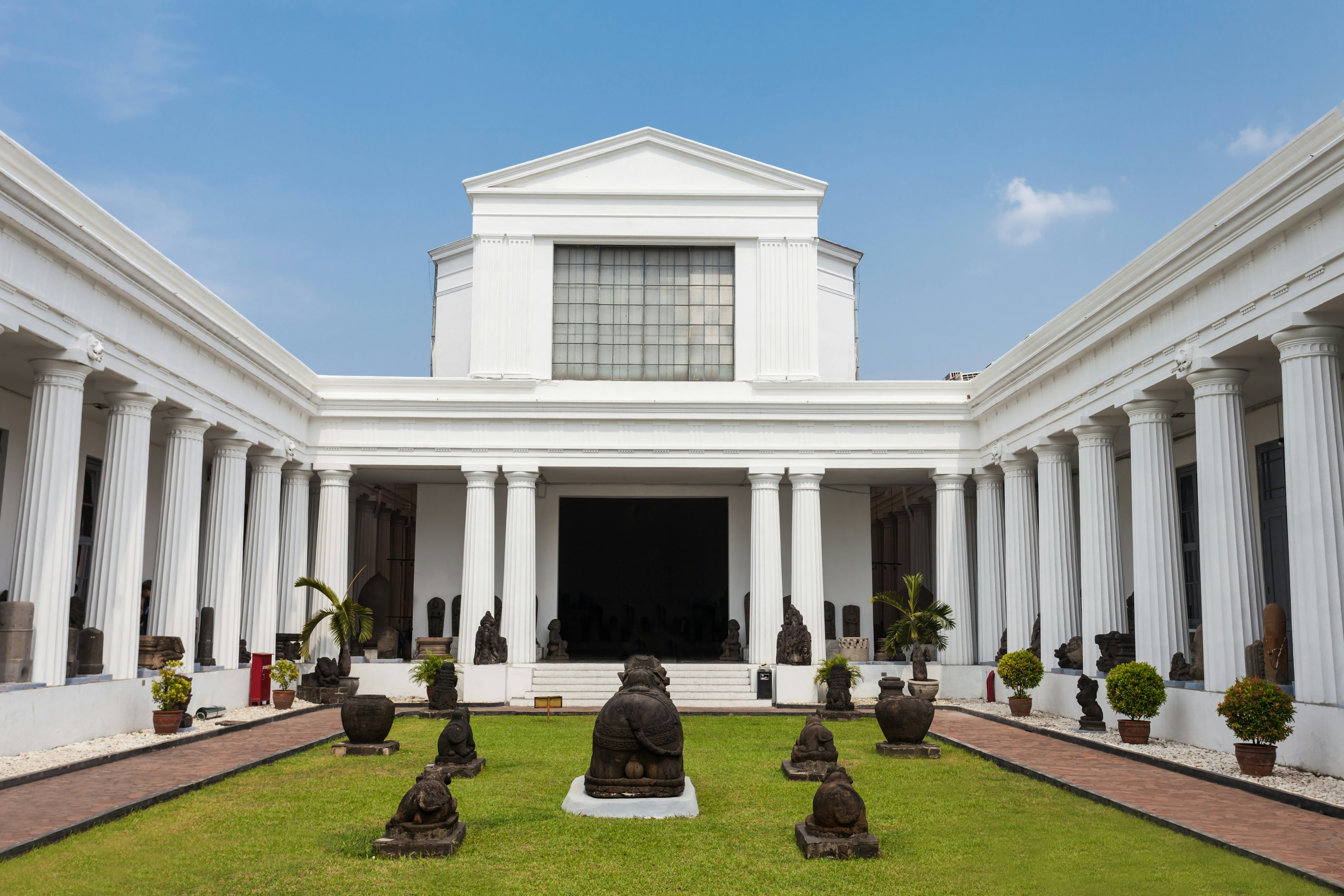 A grass courtyard with stone statues. A white building surrounds three sides; it has columns, and the central portion has a peaked second-floor tower.
