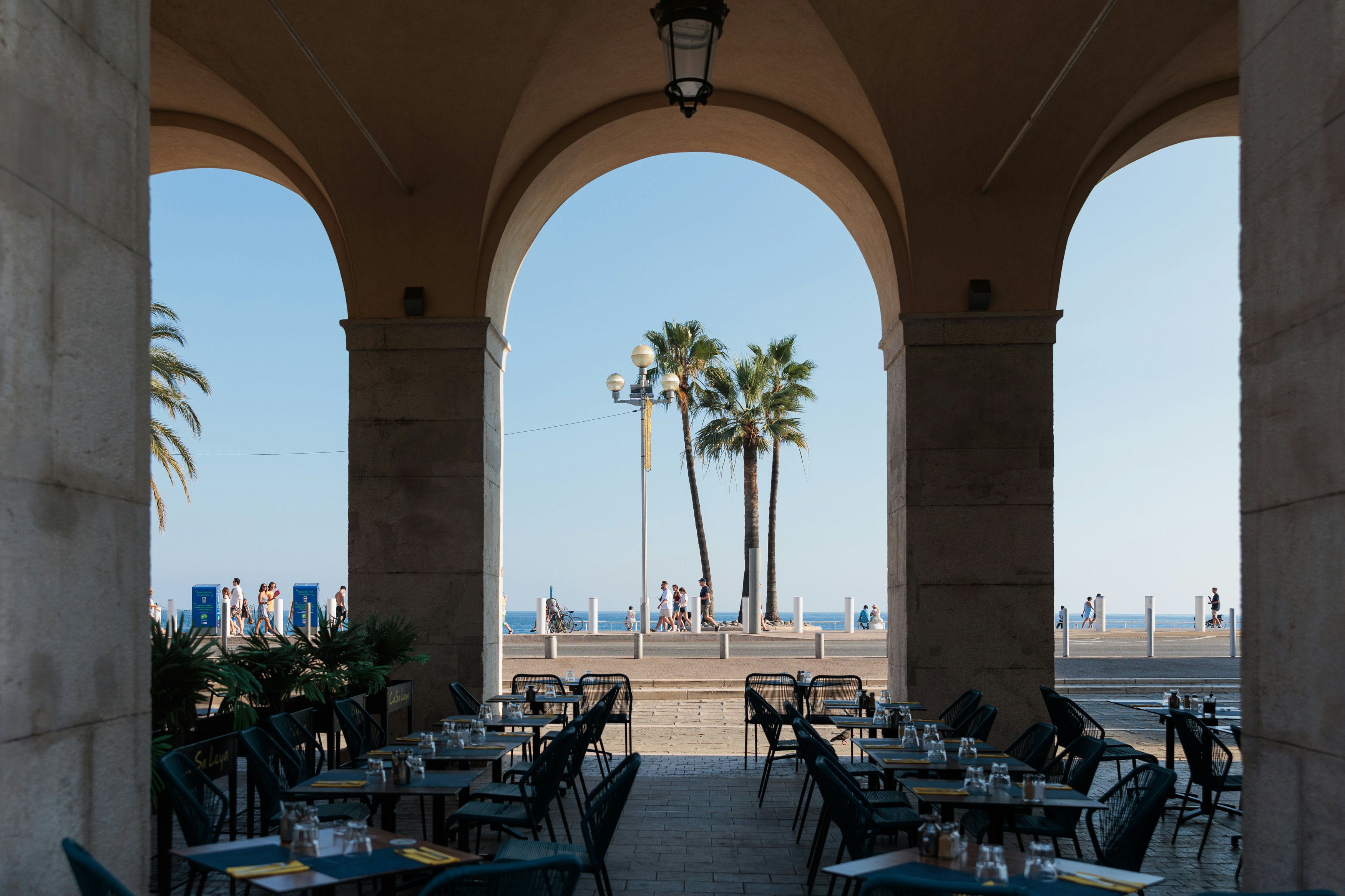 Seating and place settings on a covered terrace. People walking by and a few palm trees are in the background.