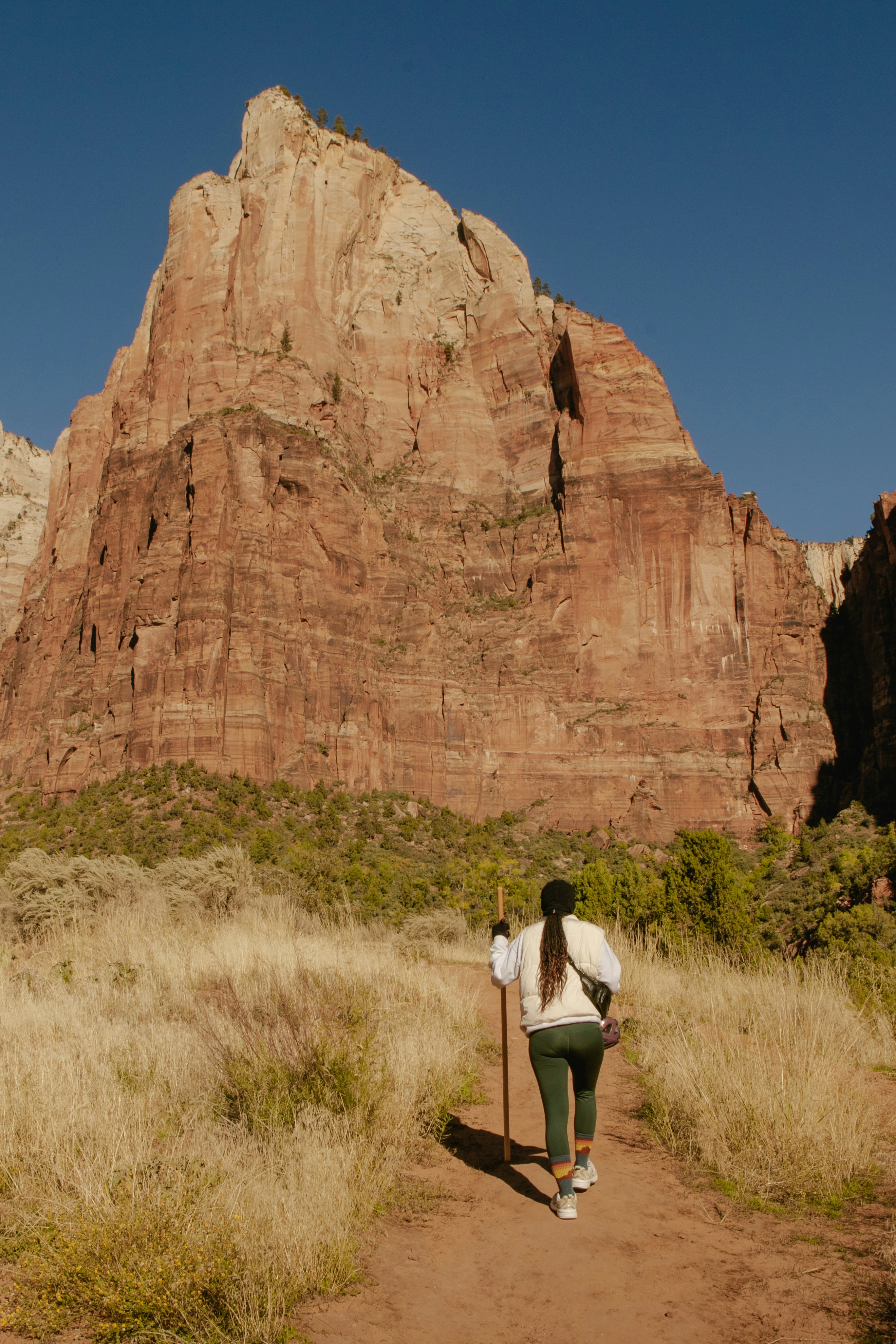 A woman with a tall stick walks on a trail toward a rocky cliff.