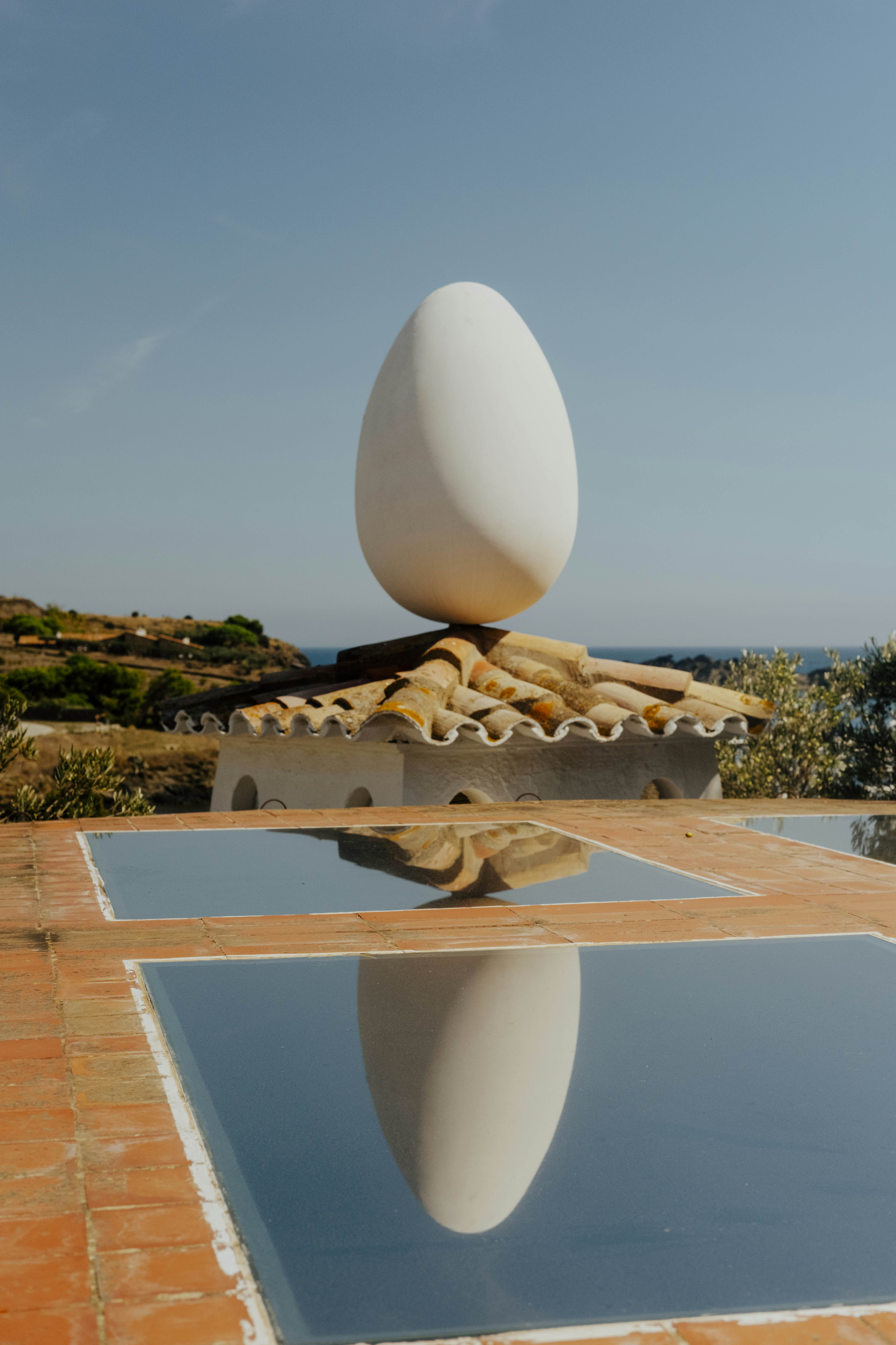 An egg-shaped sculpture balanced on a wavy roof is reflected in a pool of water.