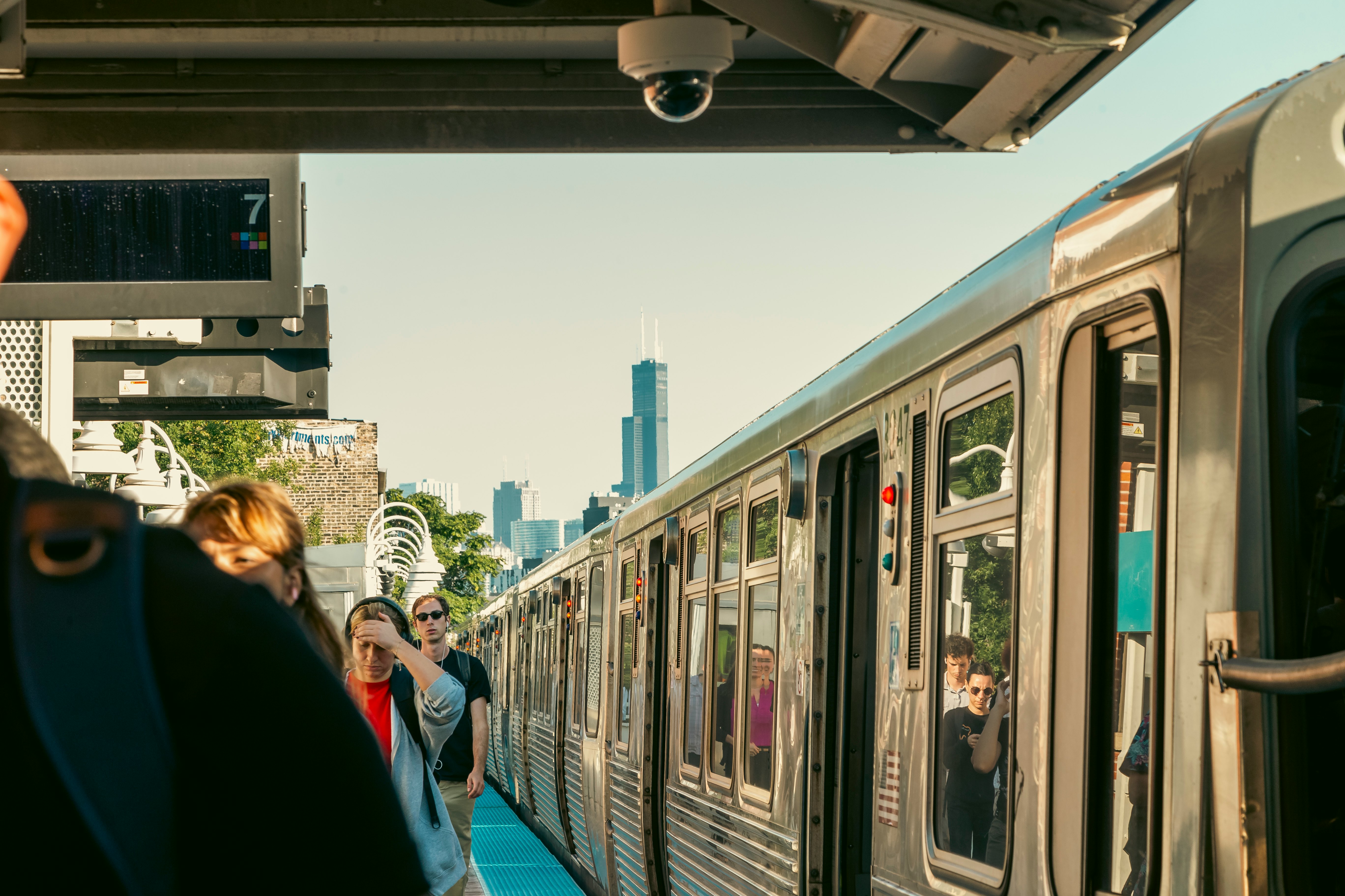 People exit a silvery train; tall towers are in the background.