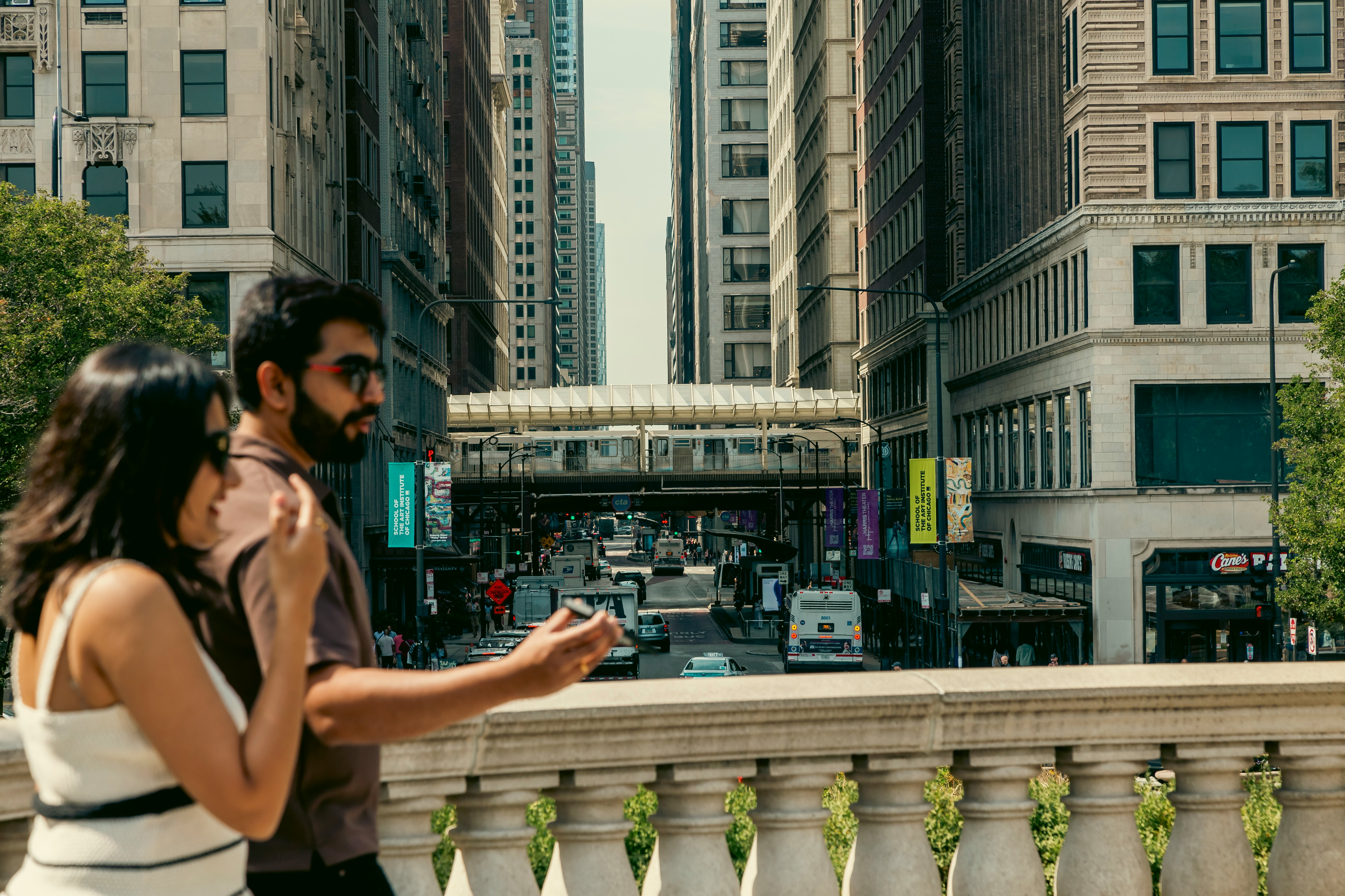 Two people walk by a stone railing; a city street runs below them, and a train station crosses over it.