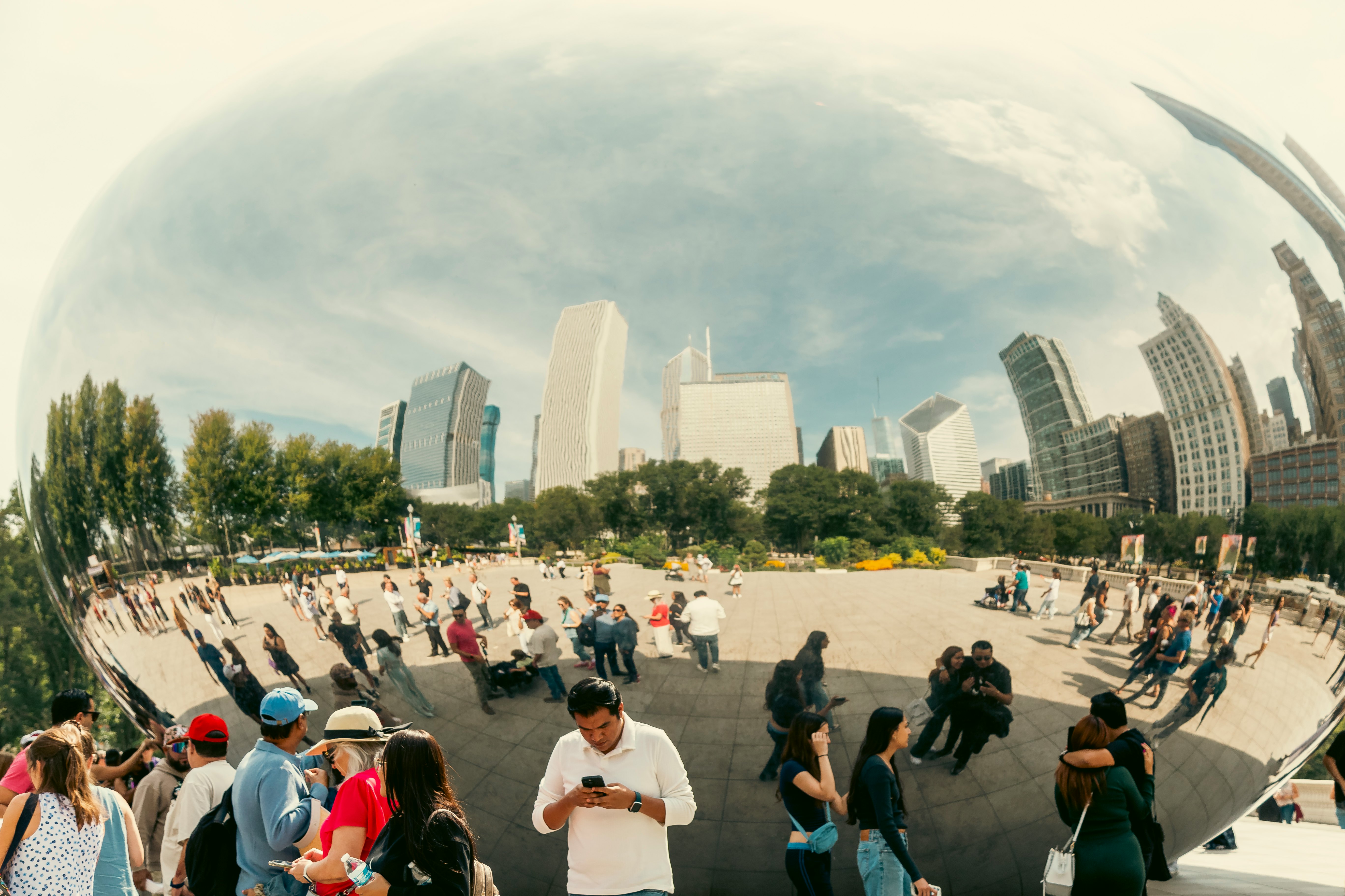 People stand in front of a large mirrored sculpture; buildings and the people are reflected on its surface.