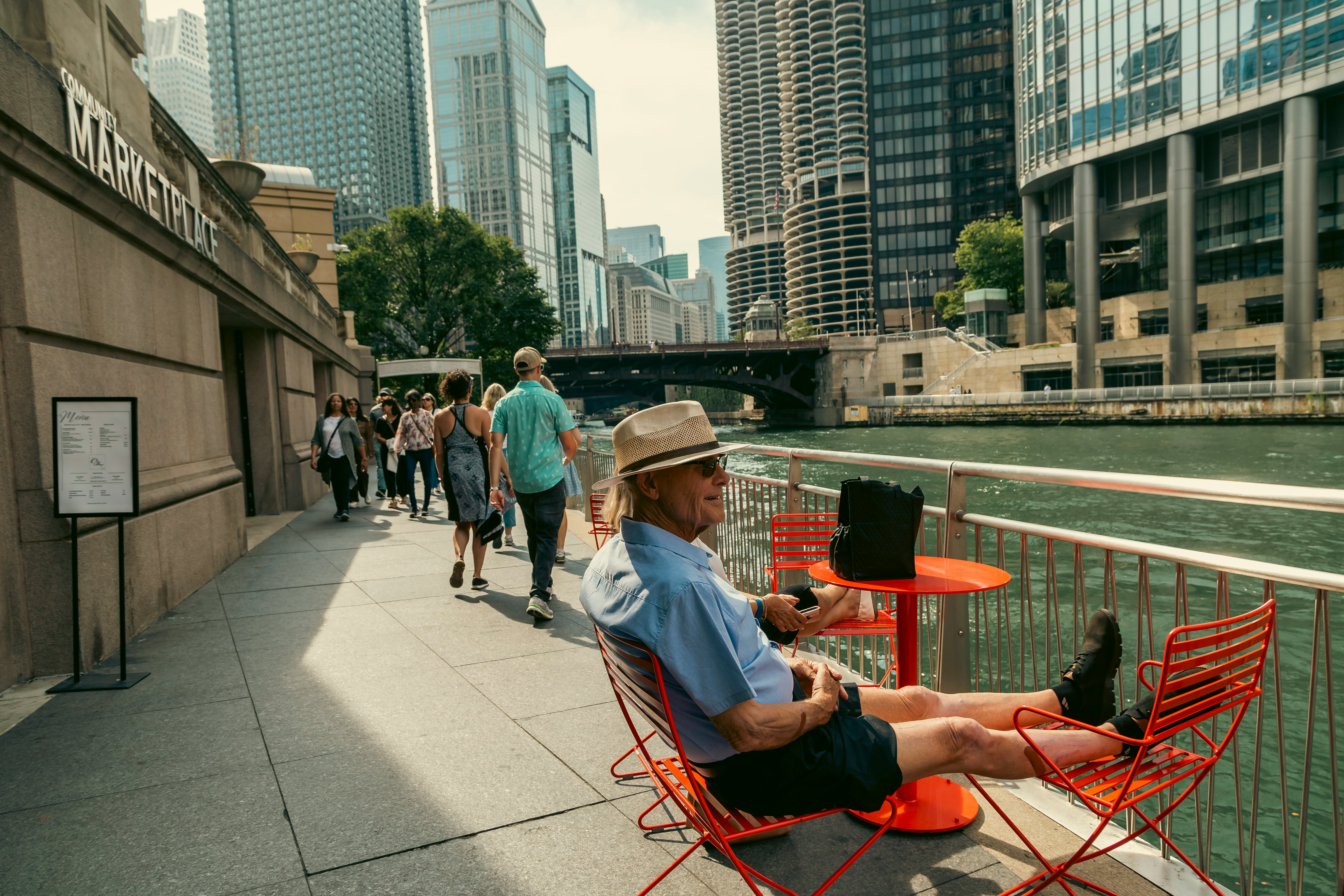 A man sits and puts his feet up on red chairs placed on a walkway along a river in a major city.