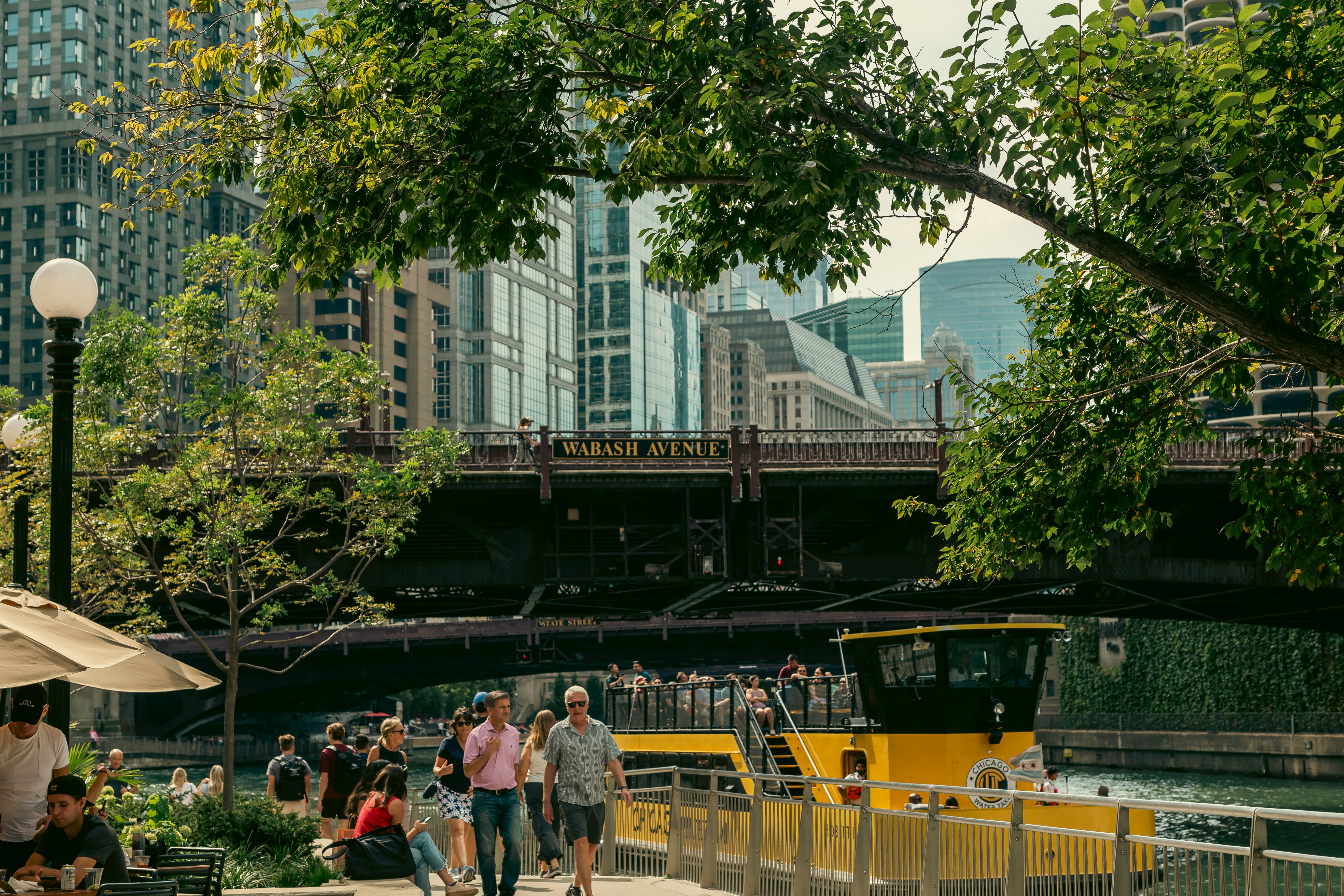 People walk by a river; there is a yellow ferry in the water and a bridge behind them with a sign that reads "Wabash Avenue."