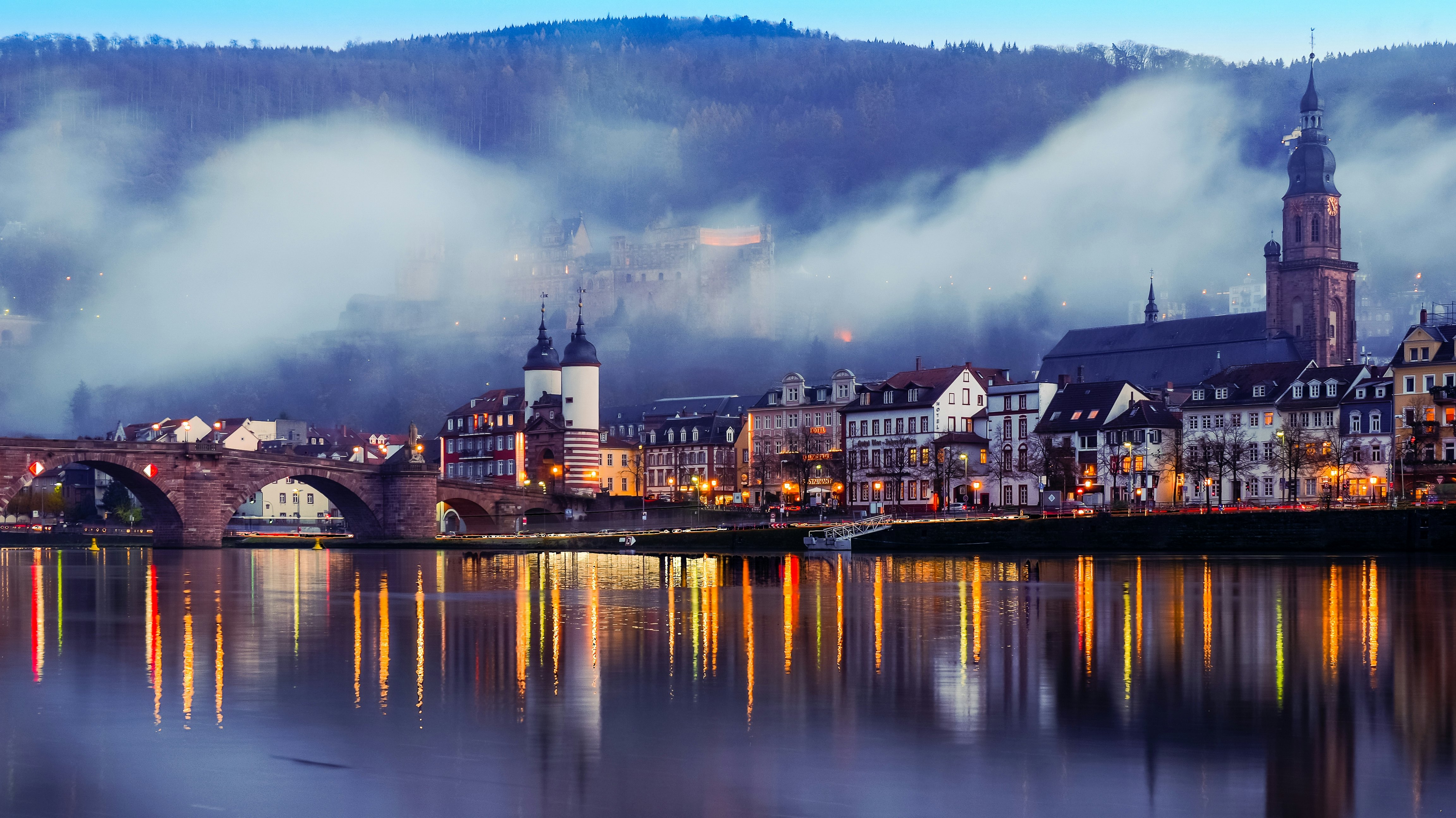 A historic city is seen at dusk, the lights of its buildings reflected in a lake, and a castle on the hill in the distance seen through mist.