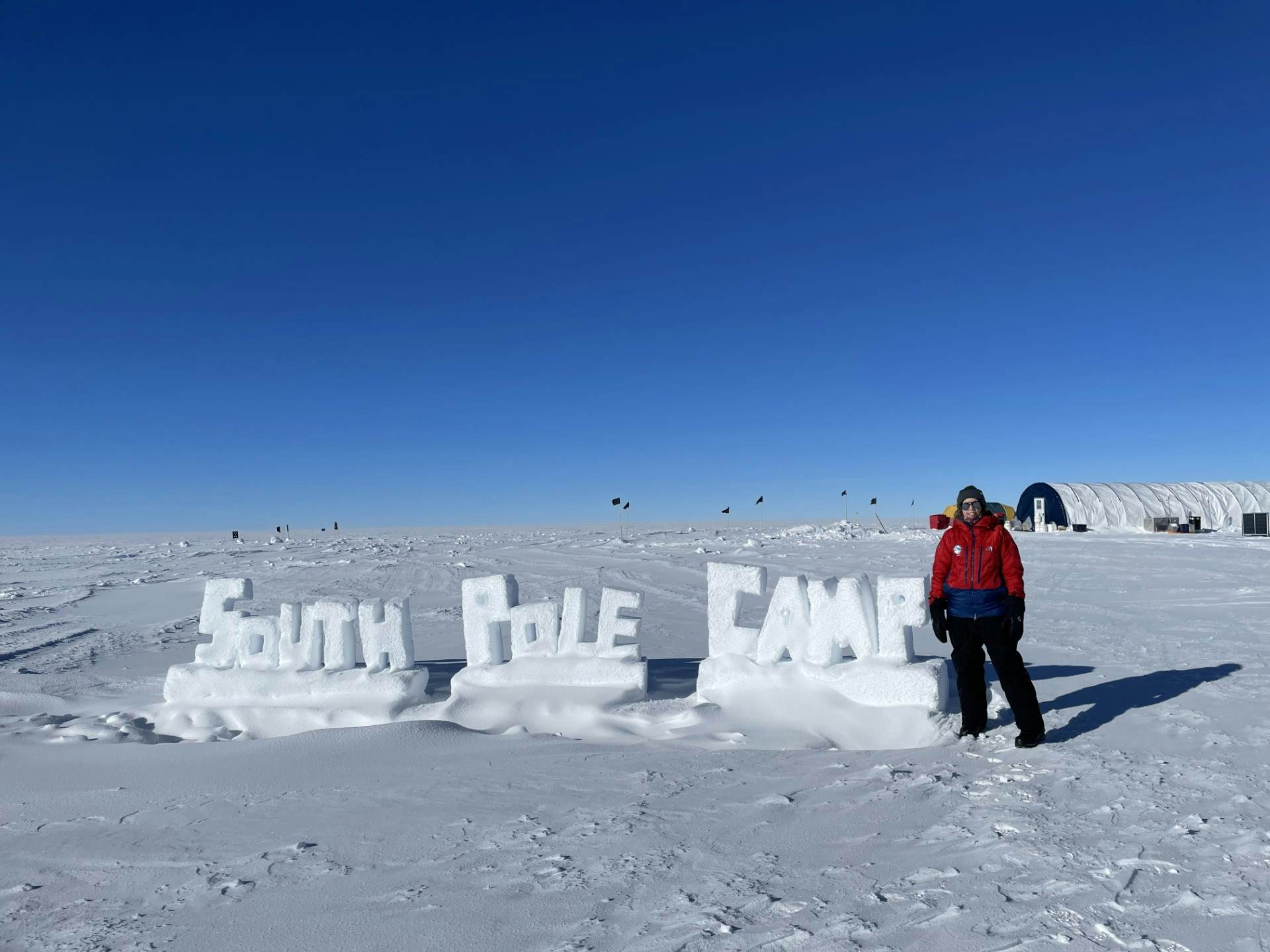 A woman stands in front of a sign that appears to be made out of snow saying "South Pole Camp"
