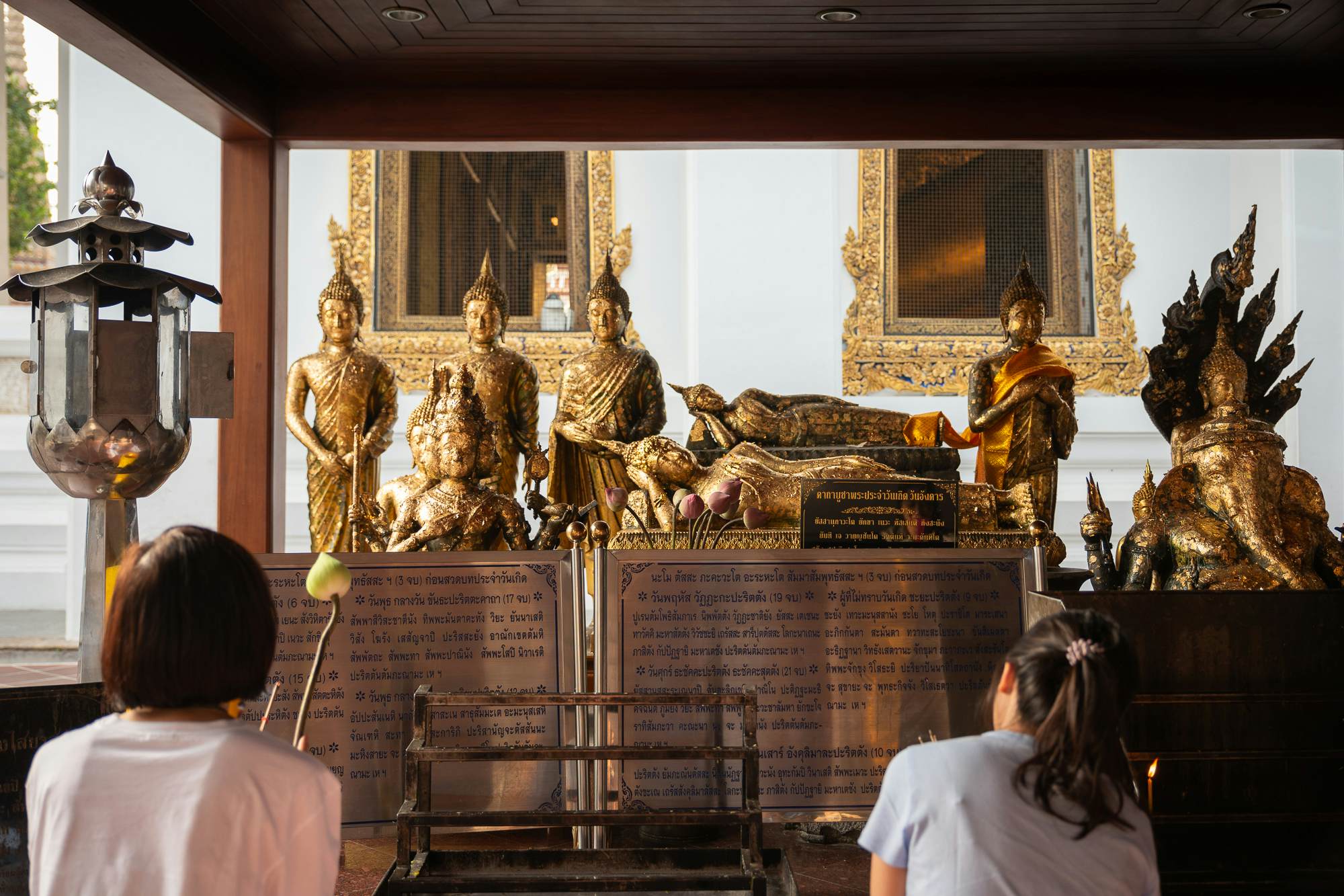 Statues of Buddha at a temple altar with two women standing in front of it with offerings.